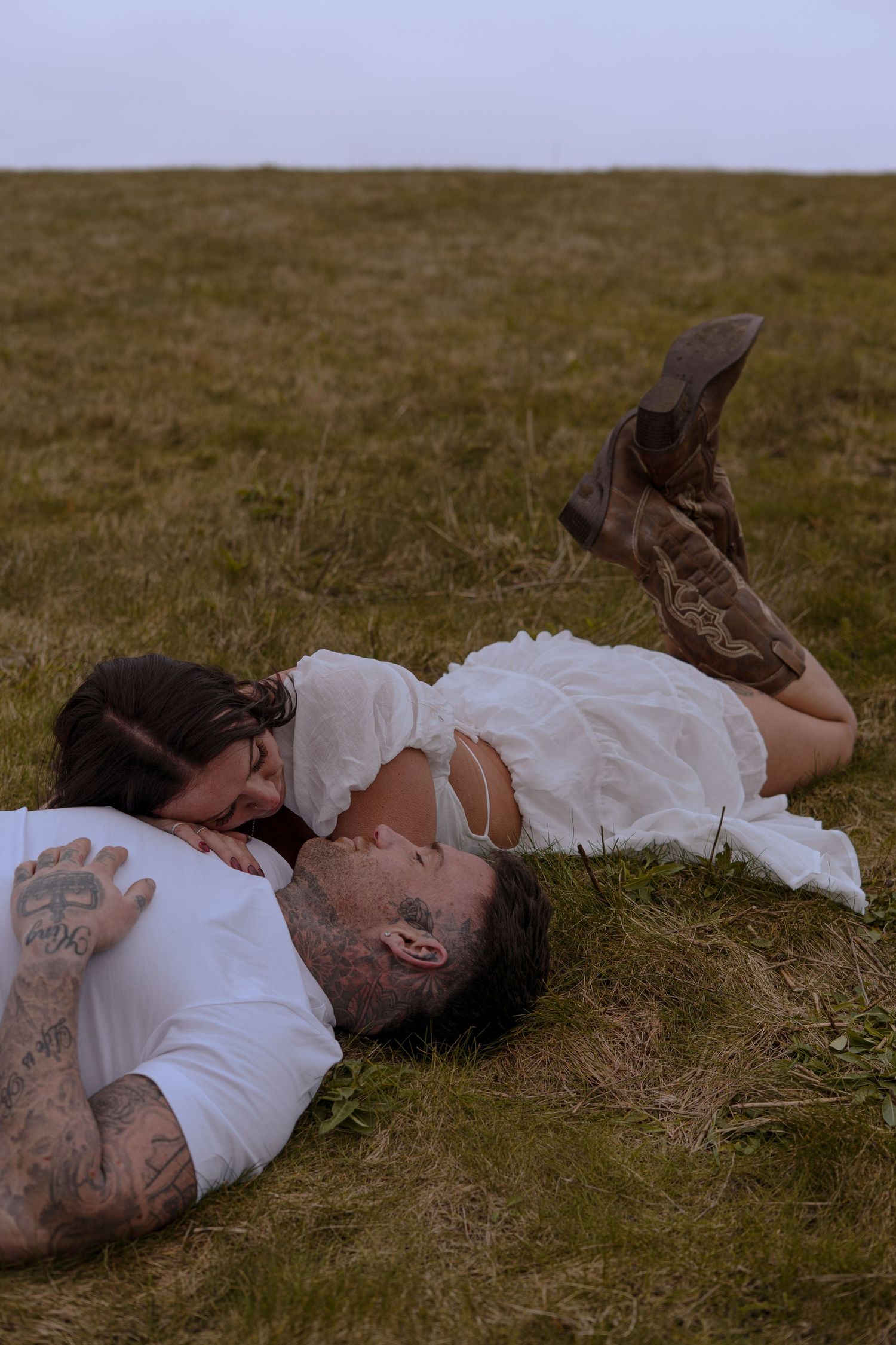 Couple lying in a grass field sharing an intimate moment, woman in white dress and cowboy boots.