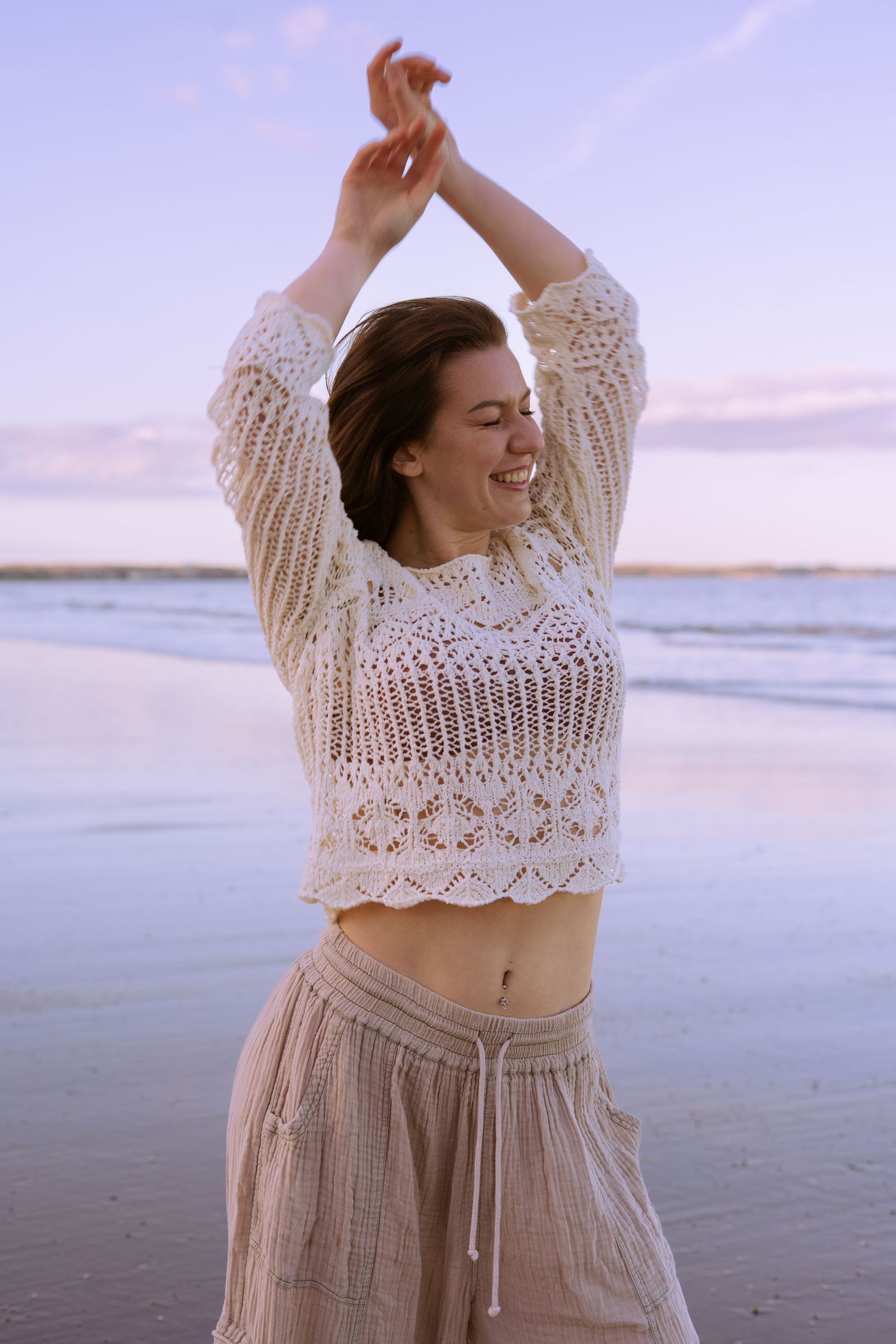 Joyful woman in crochet crop top dancing on a beach at sunset with arms raised.