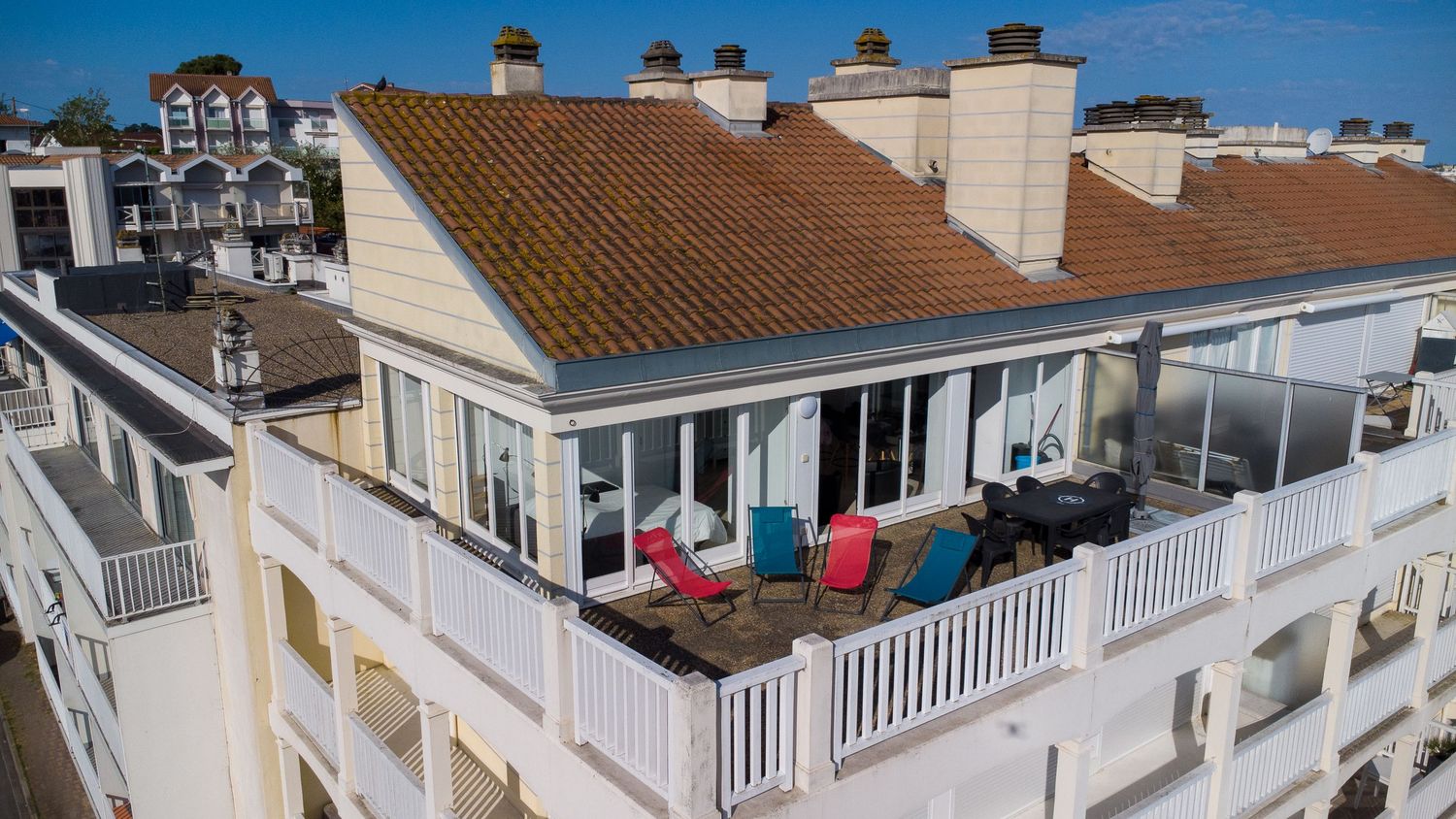 Aerial view of a luxury rooftop terrace with colorful chairs, white railings, and terracotta tile roof on a coastal building.