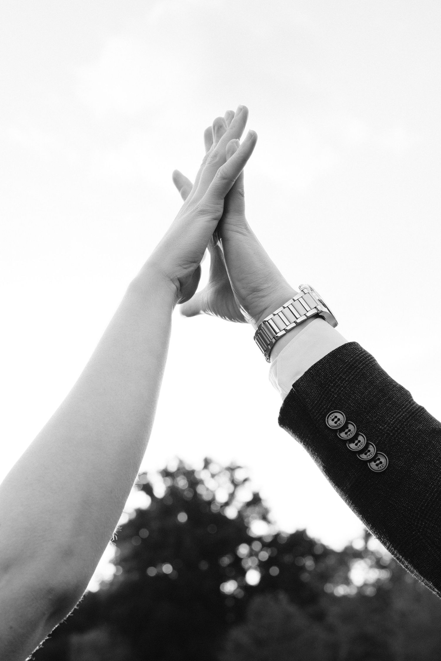 Two hands raised high giving a celebratory high five against a bright sky, one wearing a formal suit jacket and watch.
