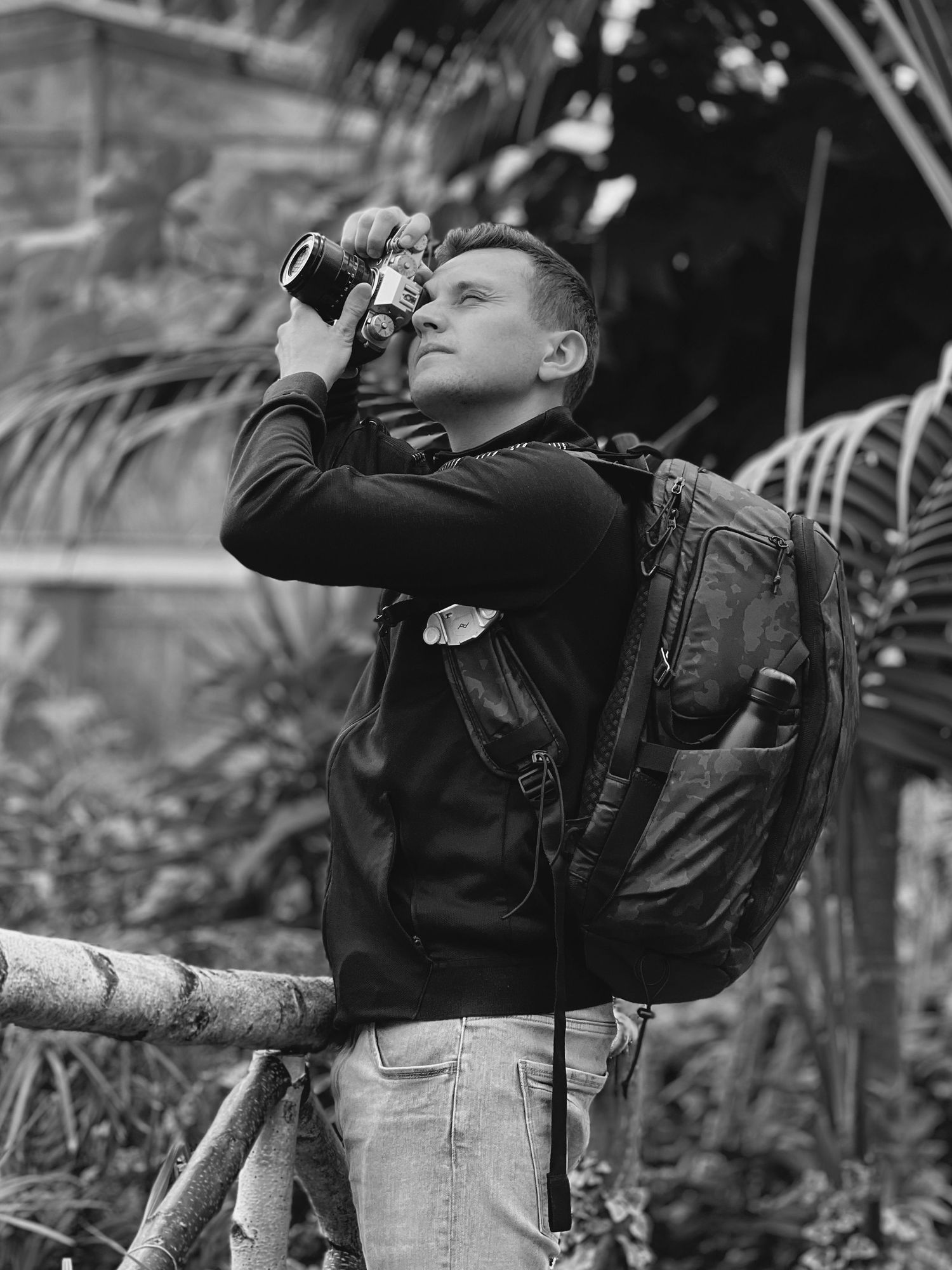 Black and white photo of a young male photographer with backpack capturing images in a tropical setting.