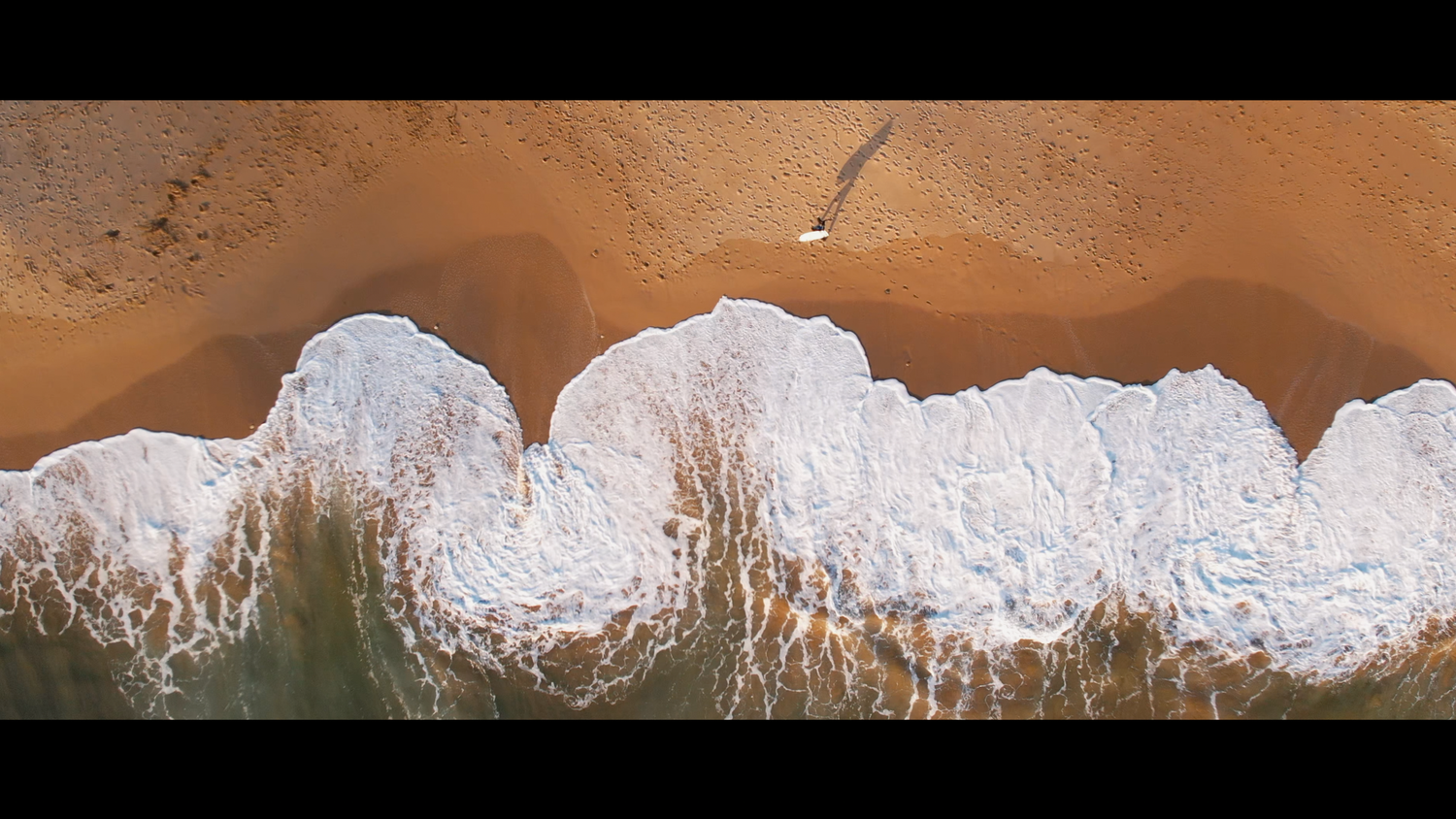 Aerial drone view of golden sandy beach with white ocean waves washing ashore in a cinematic widescreen format.