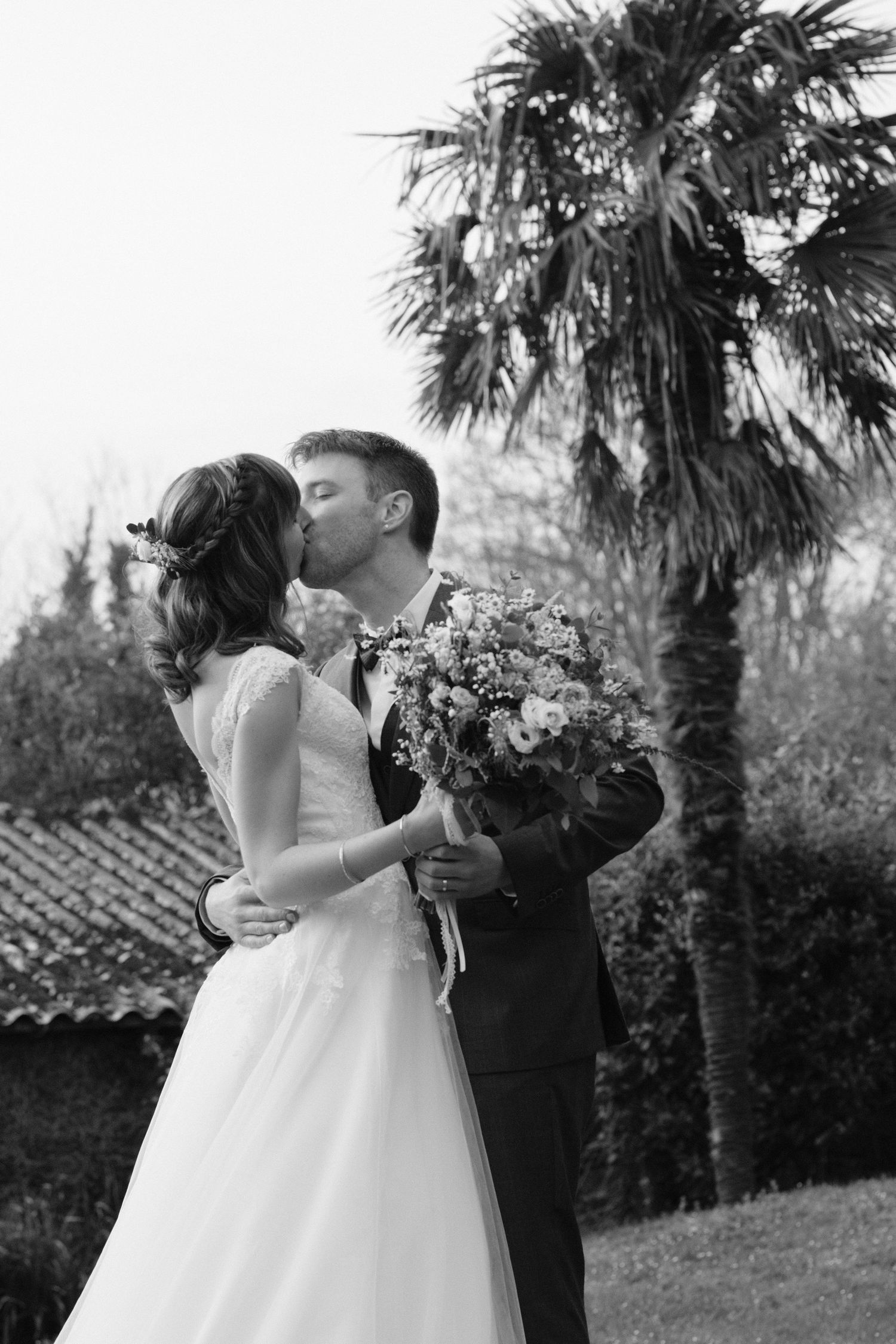 Bride and groom share a romantic kiss outdoors, with the bride holding a bouquet, palm trees in the background, black and white photo.