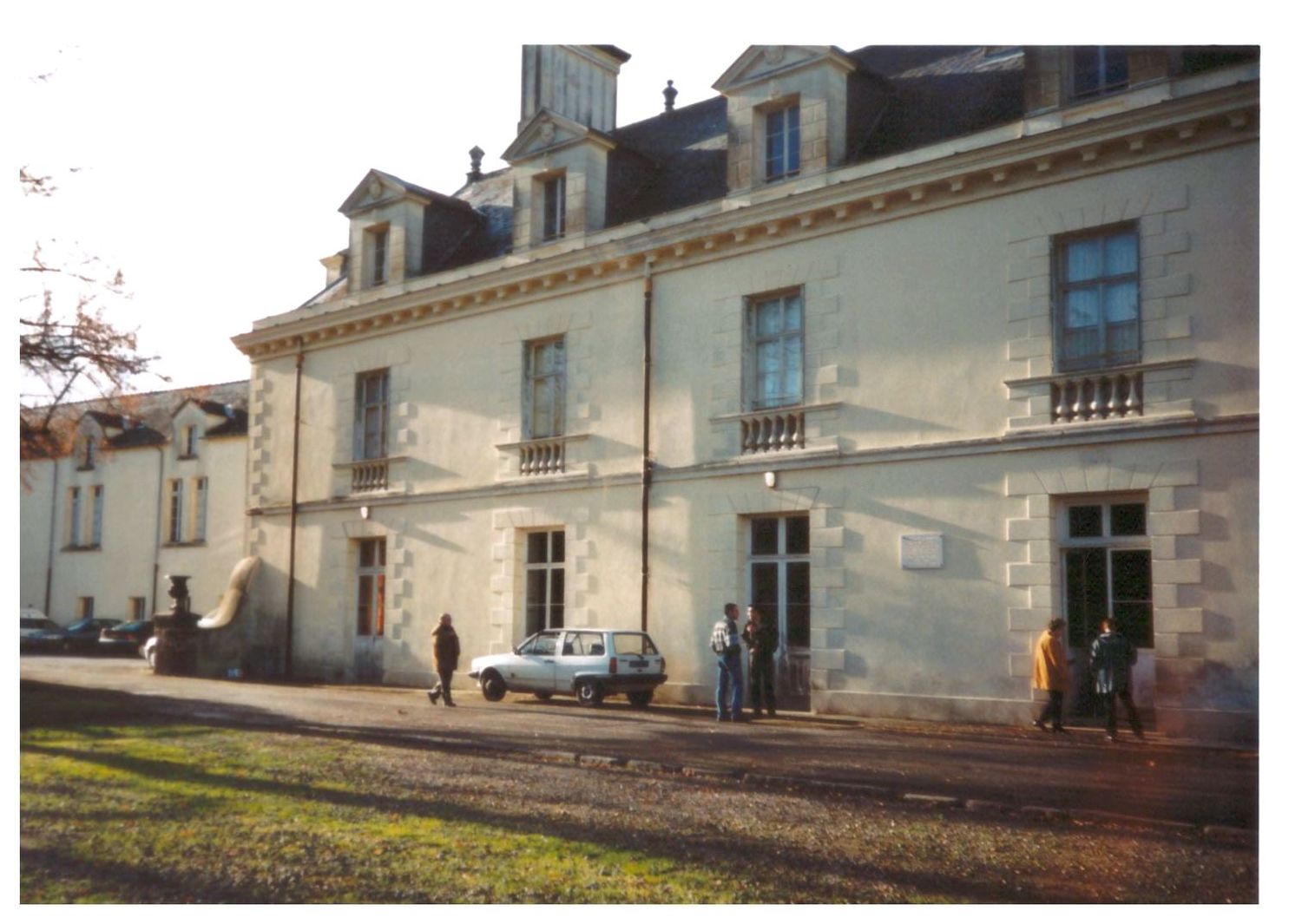 Vintage color photo of a large French manor house facade with parked cars and people outside.