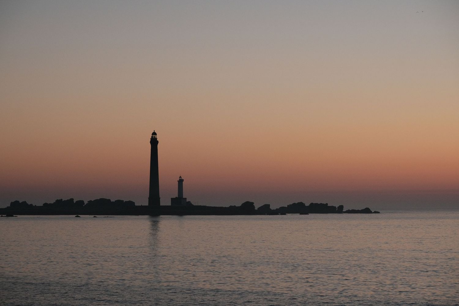 Silhouette of a tall lighthouse at sunset over calm water with warm orange and pink sky tones.