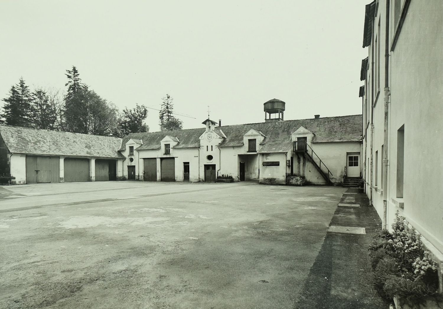 Black and white photo of a rural courtyard with outbuildings, water tower, and exterior staircase.