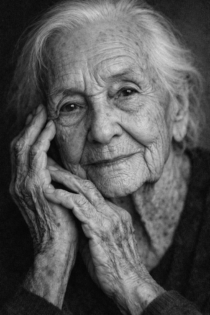 Black and white portrait of a very elderly woman resting her face in her wrinkled hands, smiling gently.