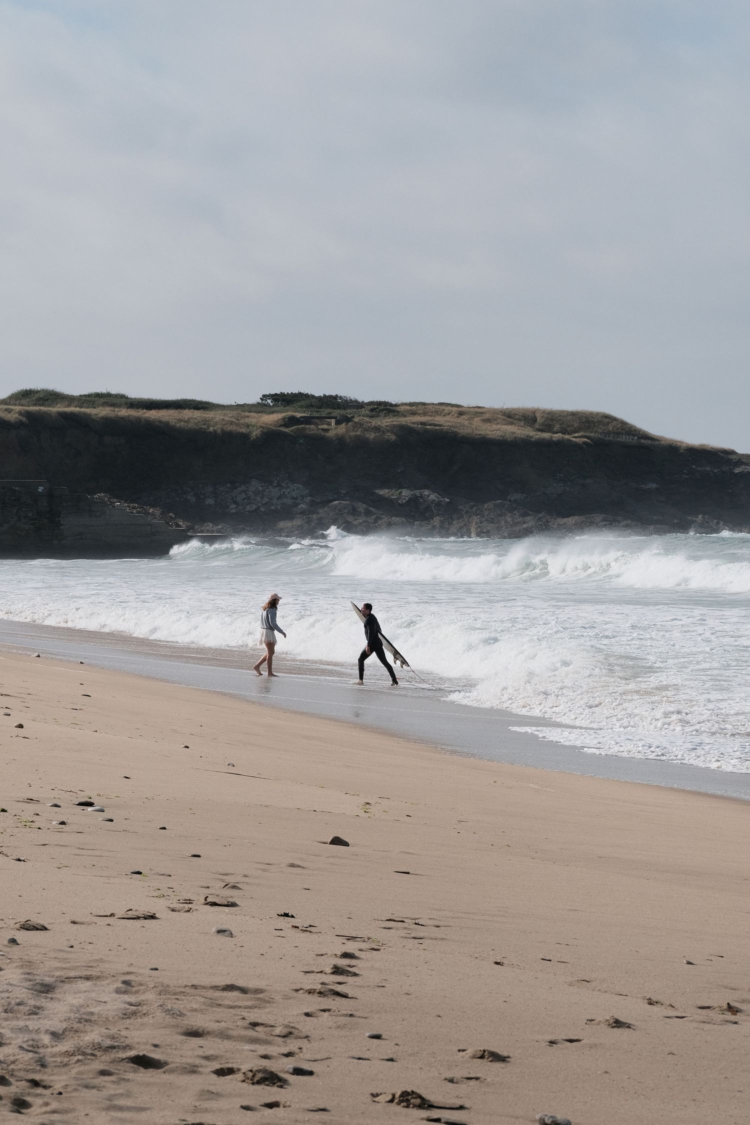 Two surfers walk along a sandy beach with crashing waves and rocky cliffs in the background on a cloudy day.