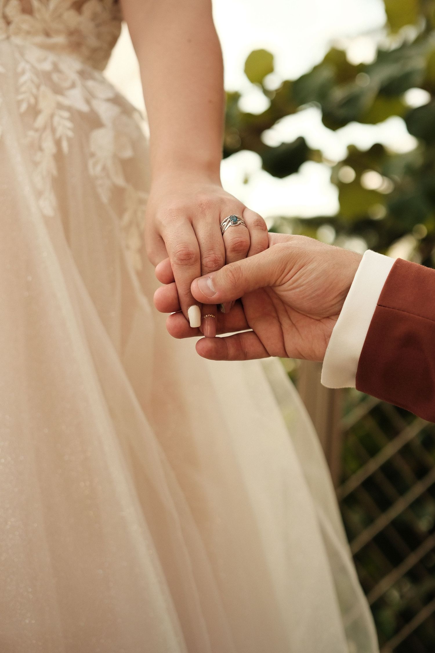 Bride and groom holding hands at wedding, bride wearing lace dress and diamond ring, groom in burgundy suit jacket.
