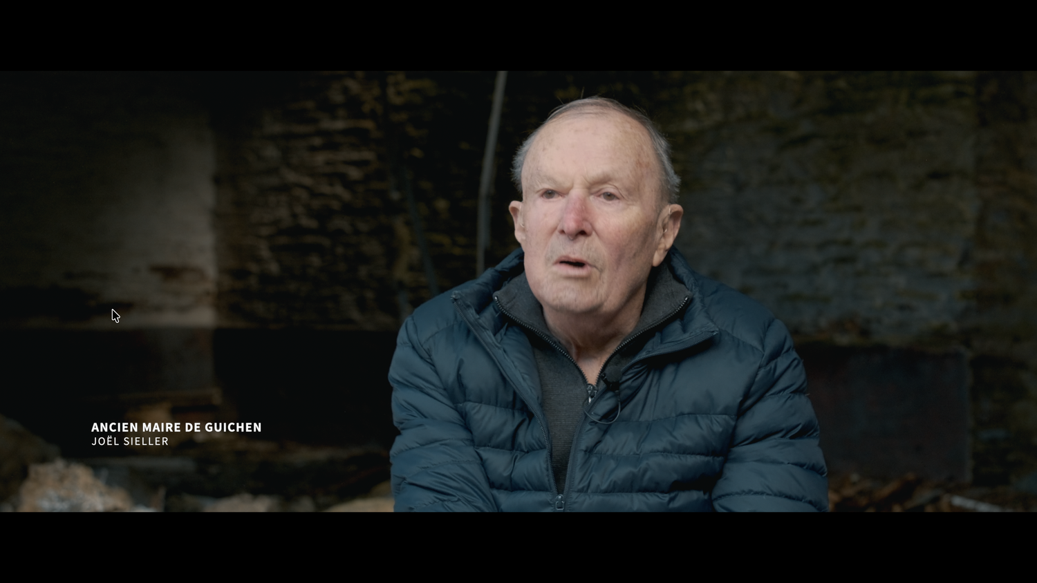 Elderly man in a blue jacket being interviewed indoors against a rustic stone wall background.