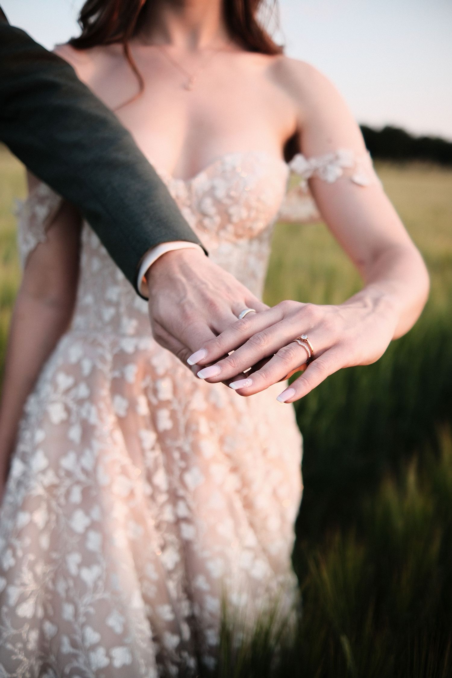 Close-up of bride and groom holding hands displaying wedding rings in a field.