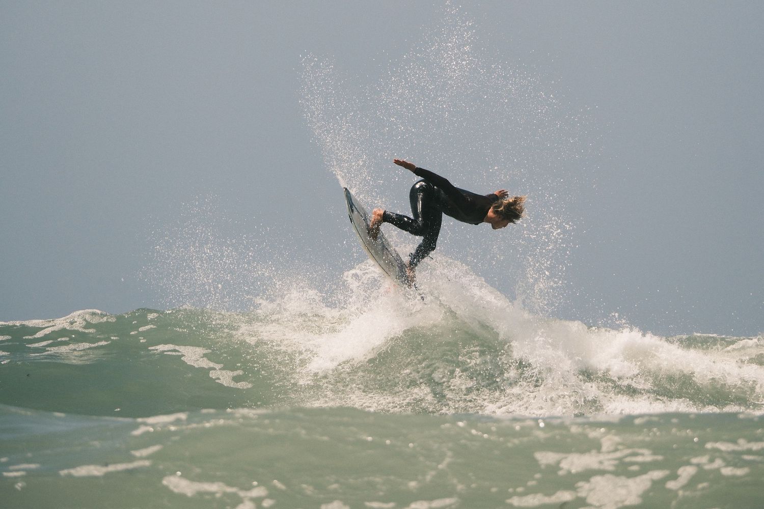 Surfer performing an aerial trick on a wave in the ocean while wearing a black wetsuit on a sunny day.