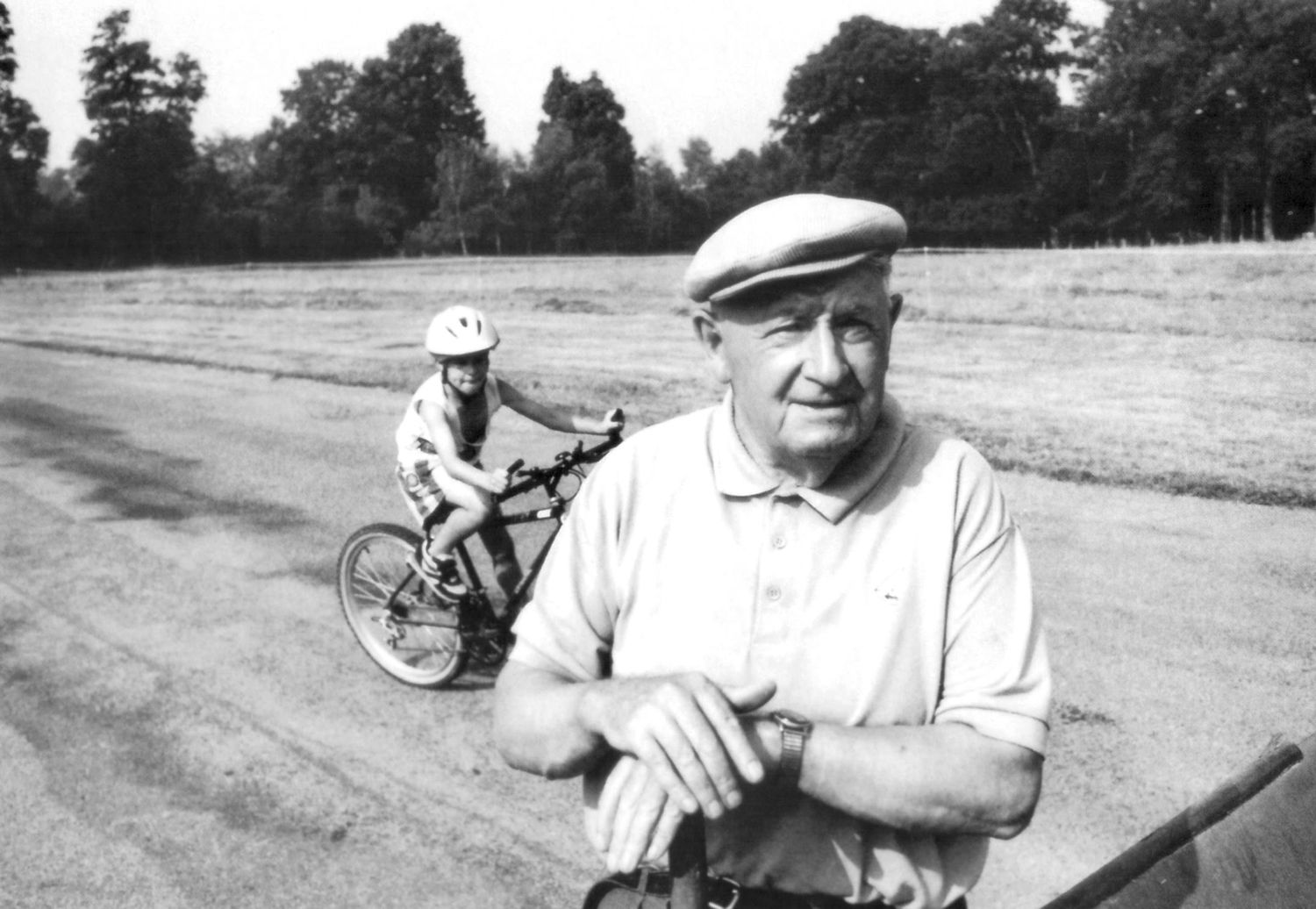 Black and white portrait of an older man in a flat cap with a cyclist on a tree-lined road behind.