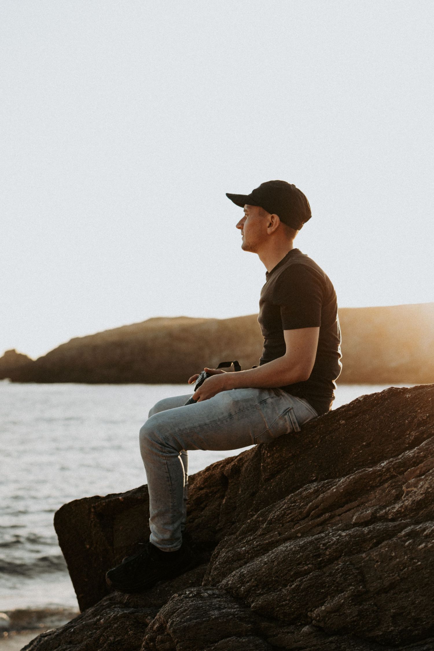 Young man wearing a black cap sitting on rocky shoreline contemplating the ocean at golden sunset.