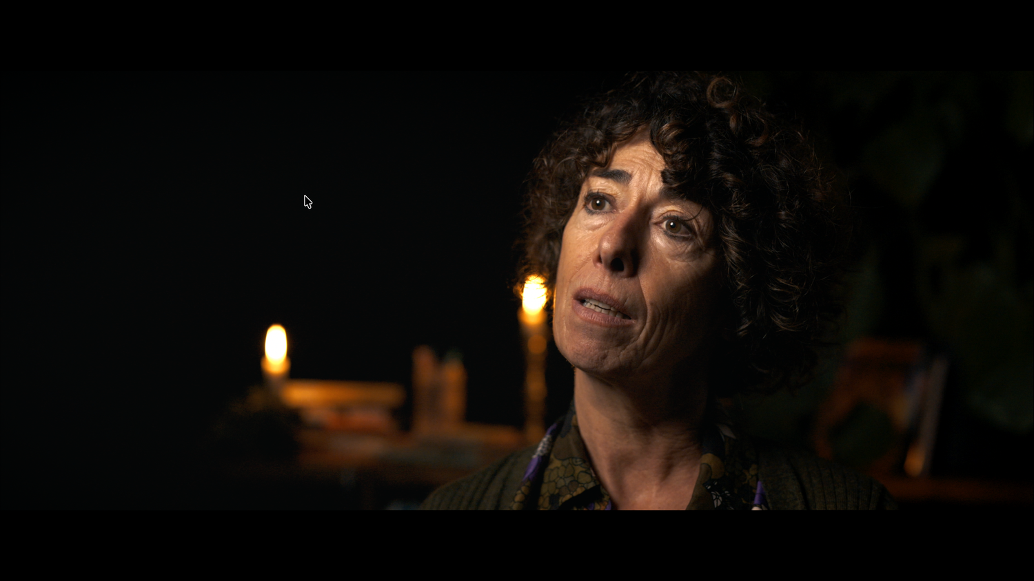A woman with curly hair gazes upward in a dimly lit scene illuminated by candlelight.