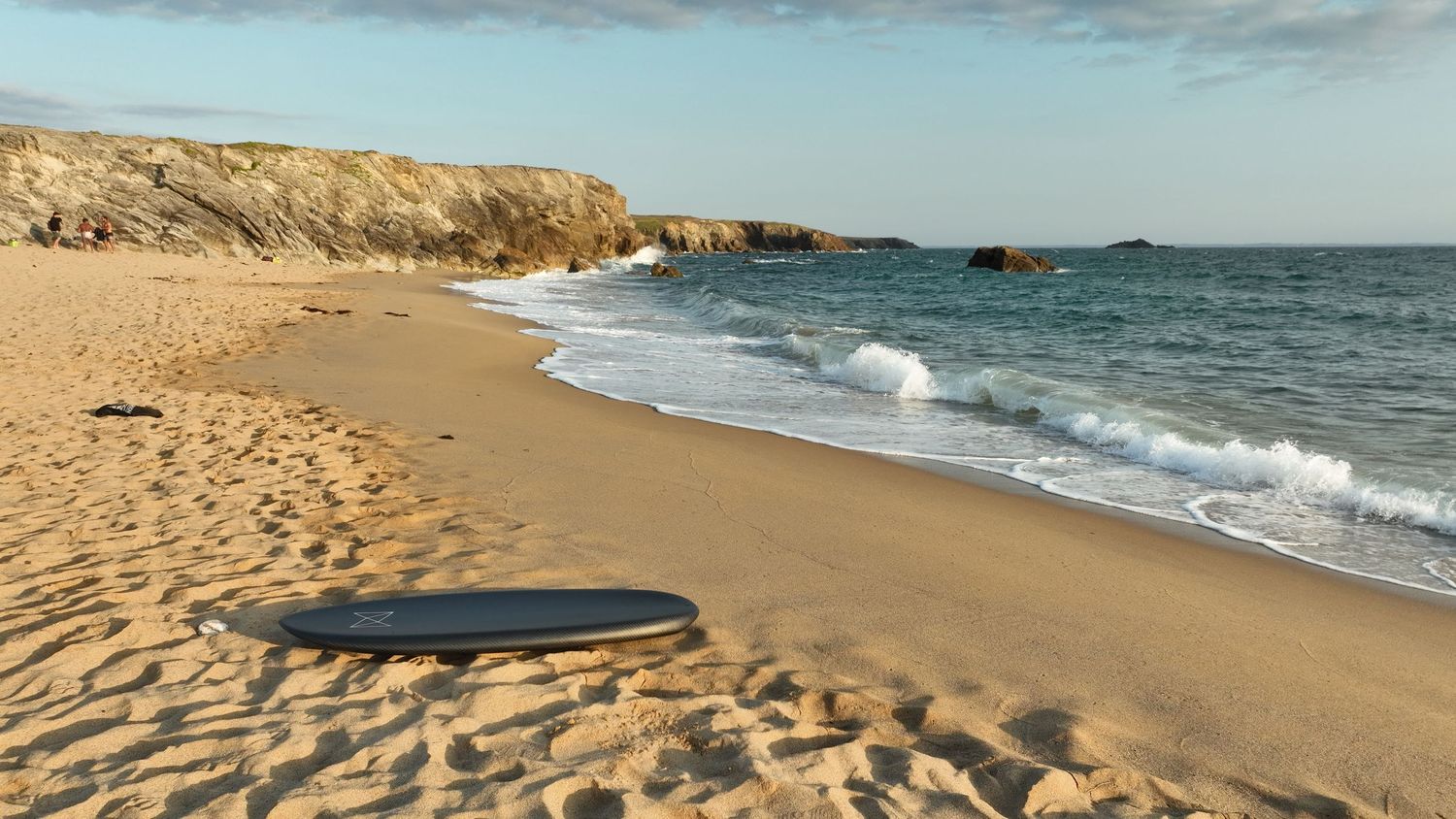 A lone surfboard rests on a golden sandy beach with rocky cliffs and gentle ocean waves under a clear sky.