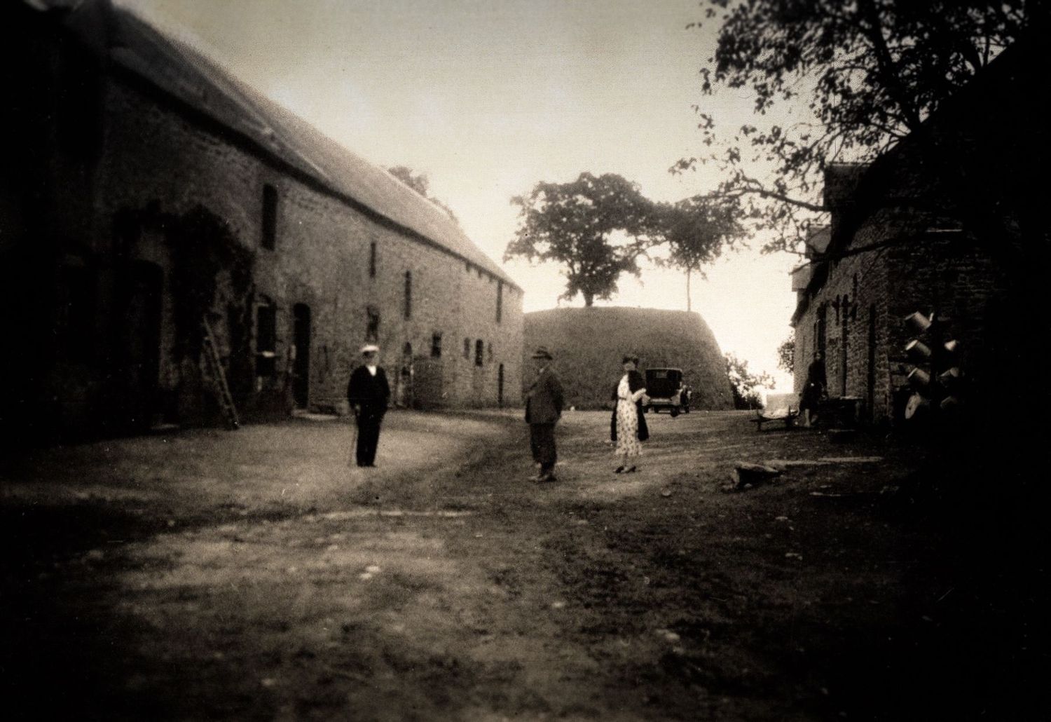 Vintage sepia photo of people walking through a rustic farm courtyard with stone buildings.