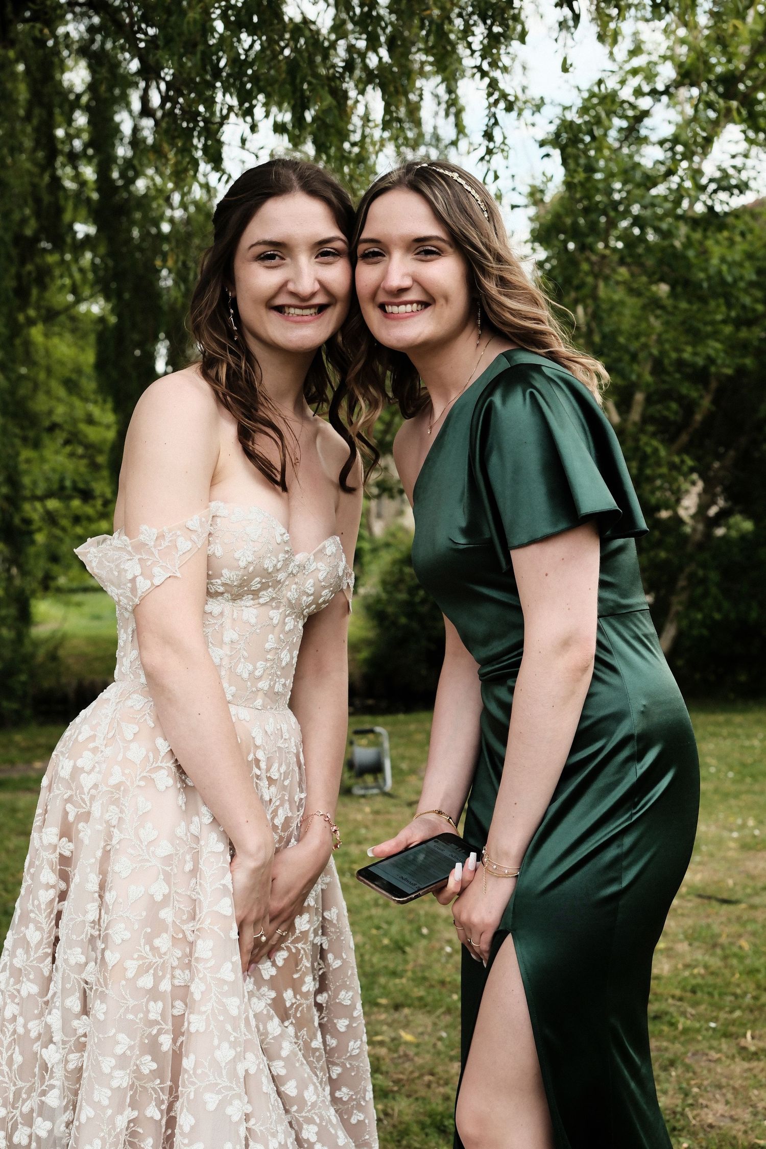 Two smiling women in formal dresses, one in floral lace and one in emerald green, pose outdoors.