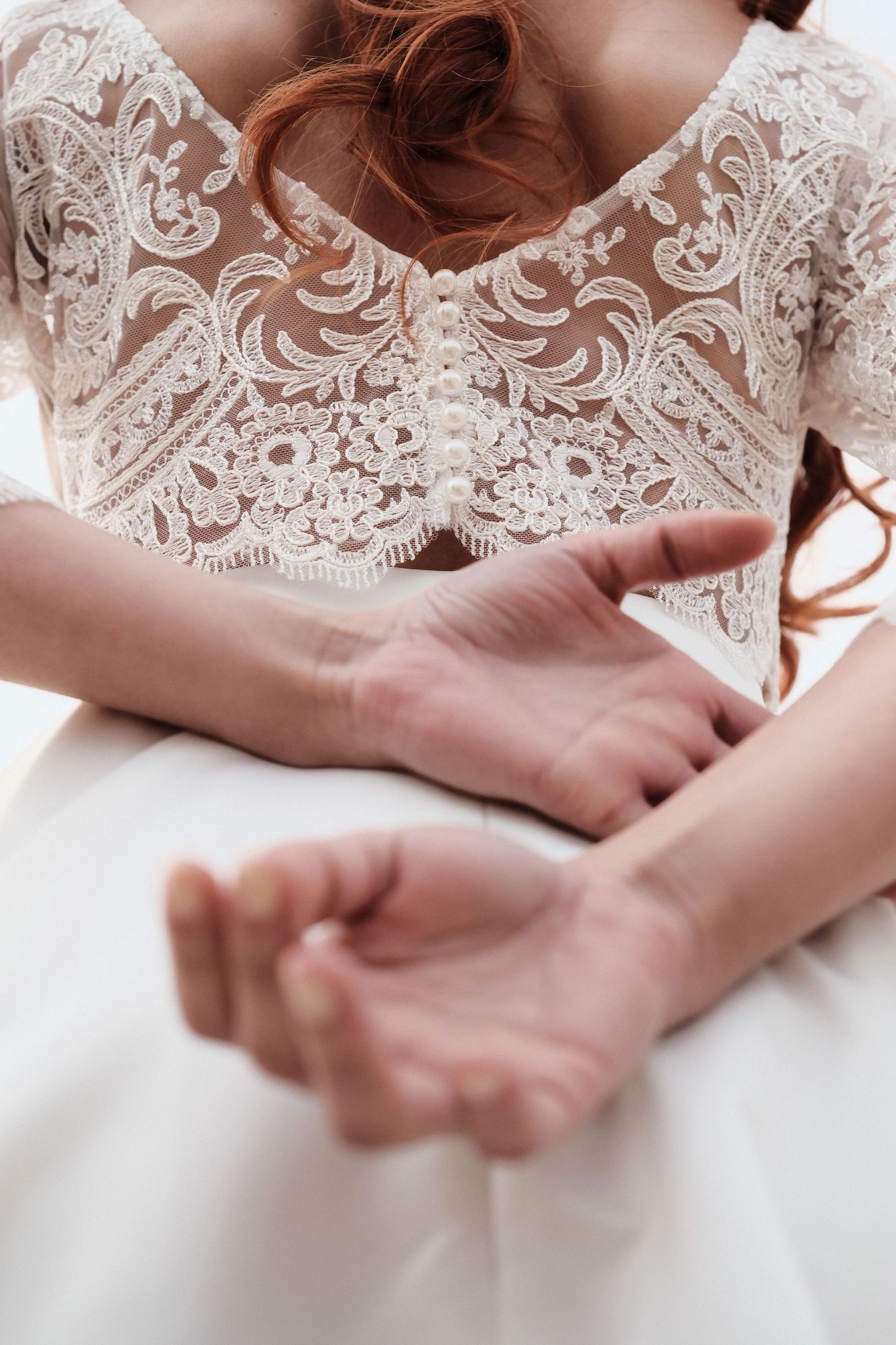 Bride in lace wedding dress with pearl buttons, hands gently crossed over white skirt.