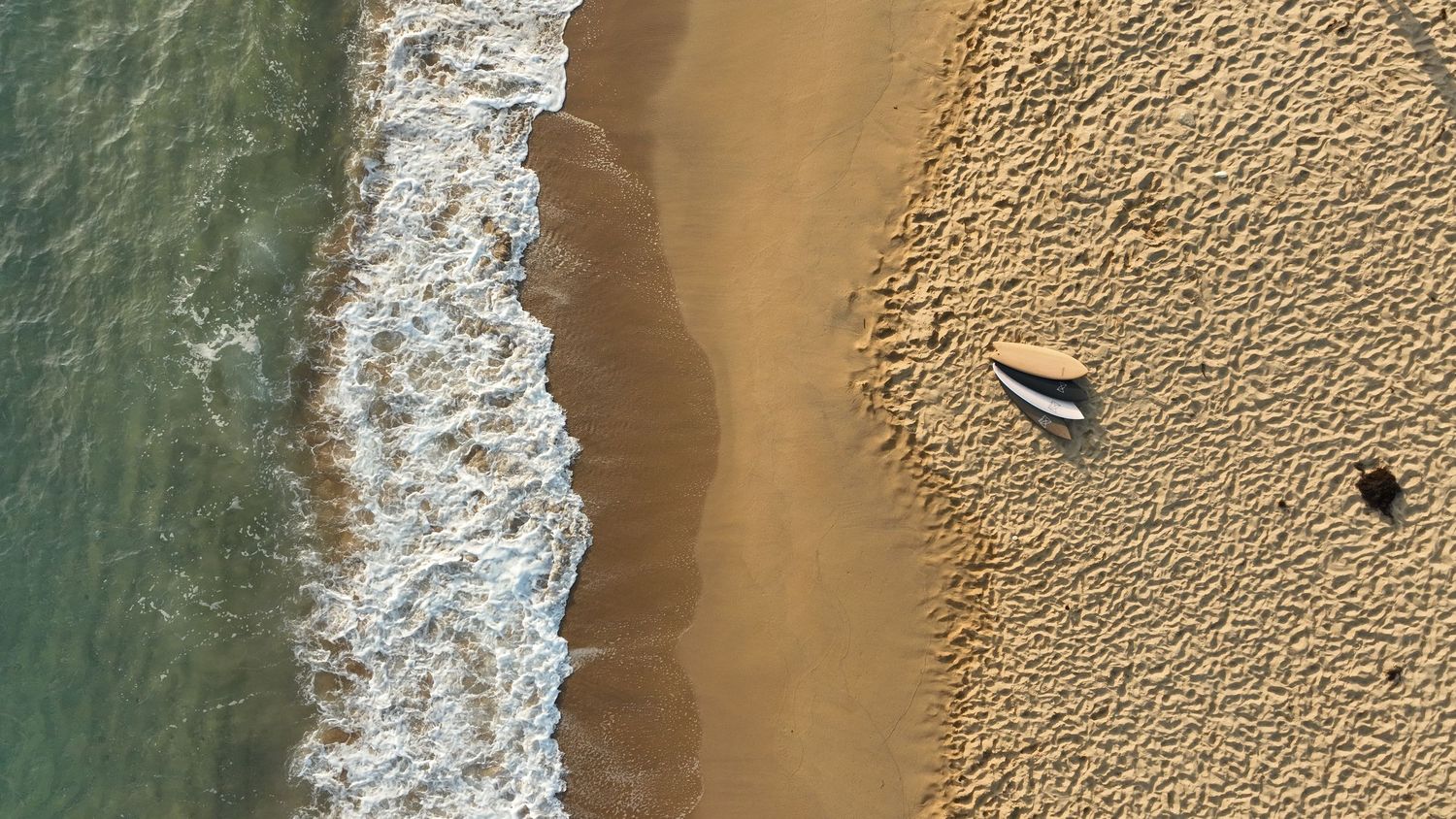 Aerial view of sandy beach with gentle ocean waves washing ashore and a small boat resting on the sand.