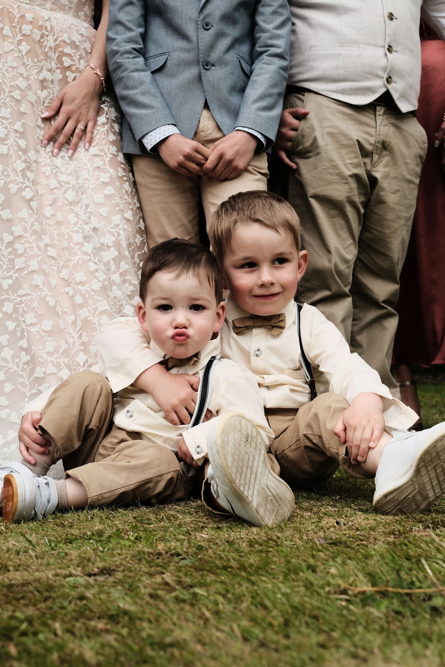 Two young ring bearers in suspenders and bow ties sit together on grass at a wedding.