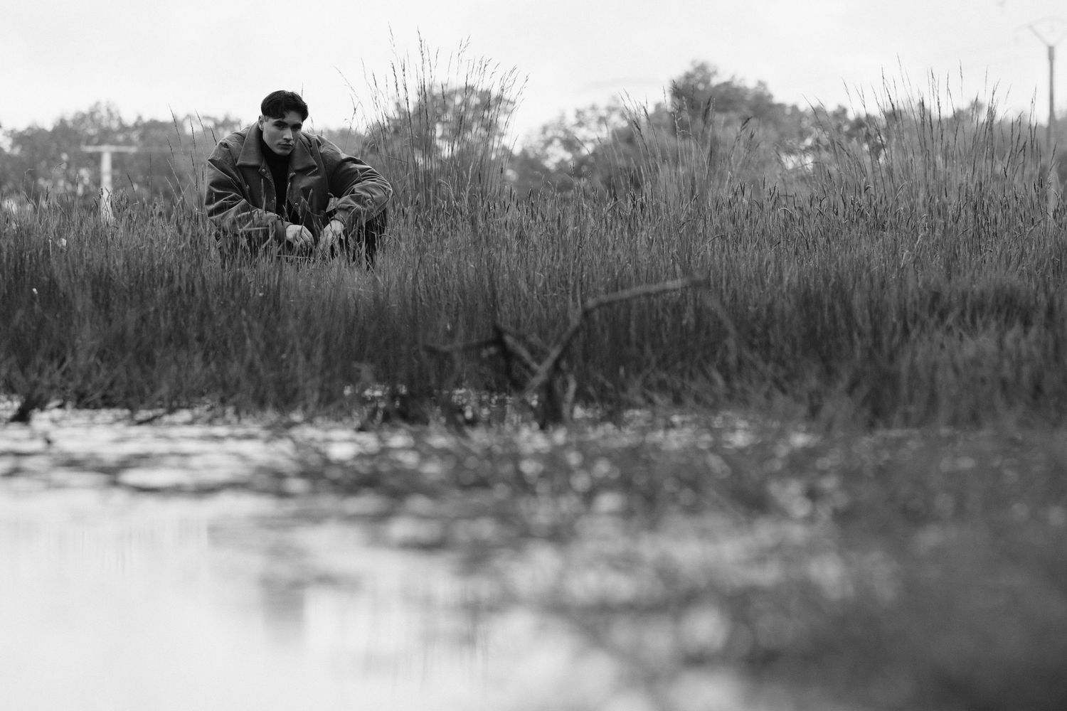 Black and white photo of a man sitting alone by a marshy wetland with tall reeds.