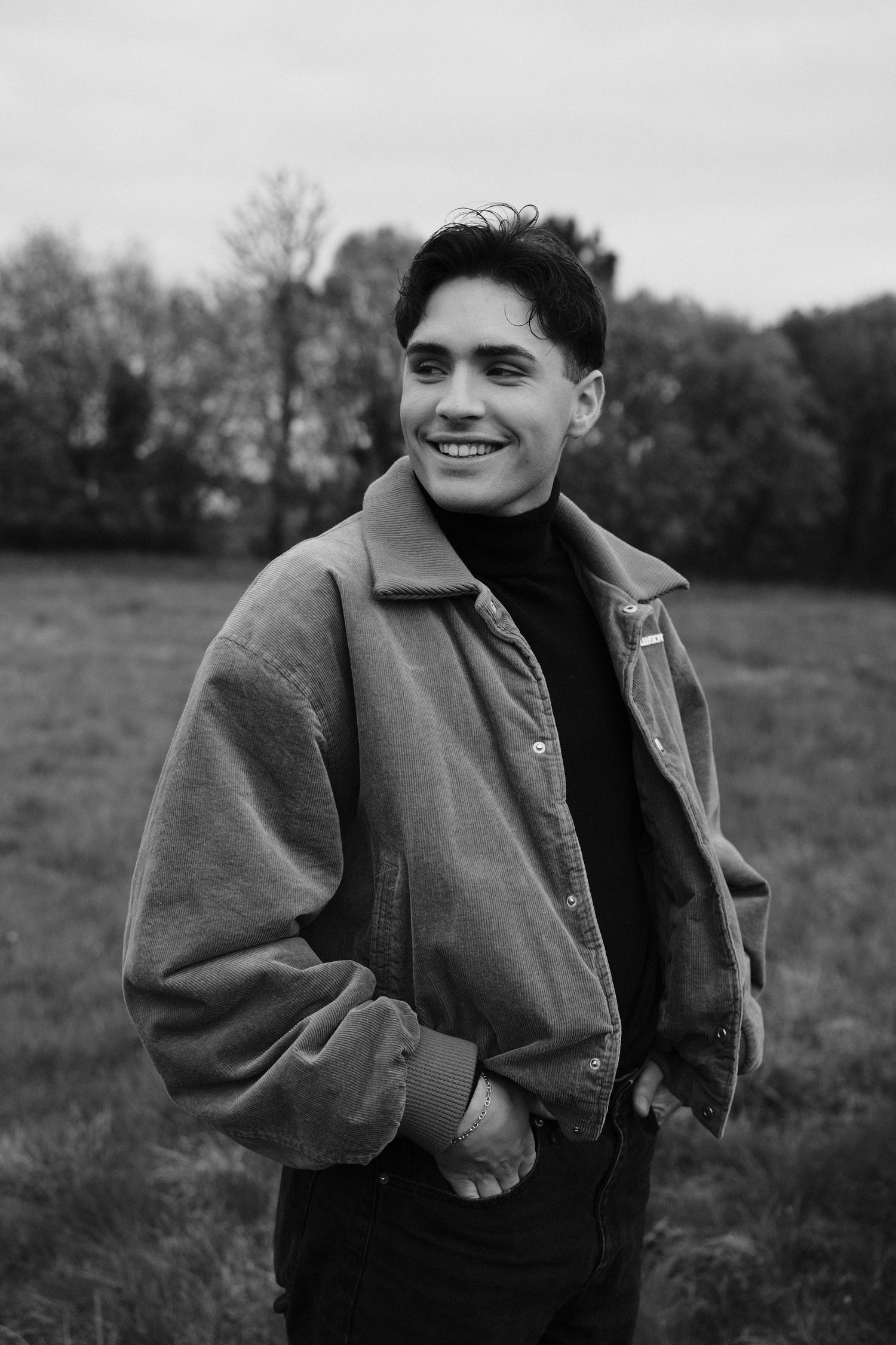 Smiling young man in a casual jacket stands in a field with hands in pockets, black and white outdoor portrait.