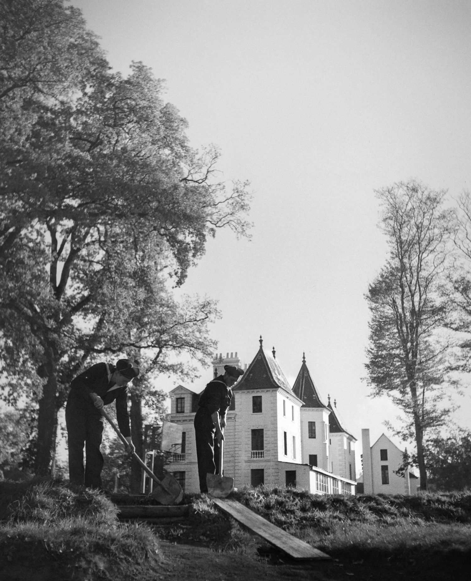 Black and white photo of two workers with tools near a chateau with turrets surrounded by trees.