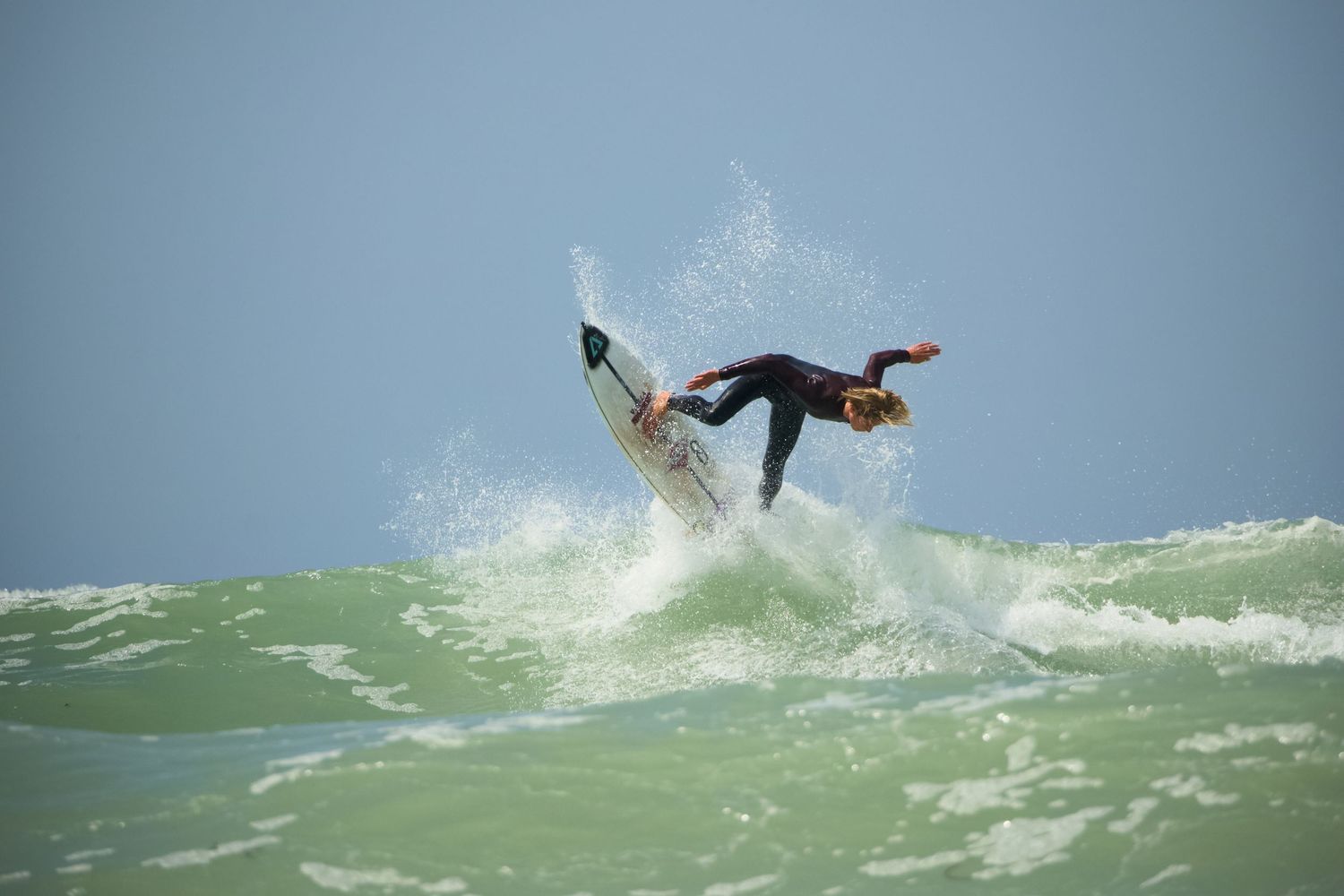 Surfer in a wetsuit performing an aerial maneuver on a green ocean wave against a clear blue sky.