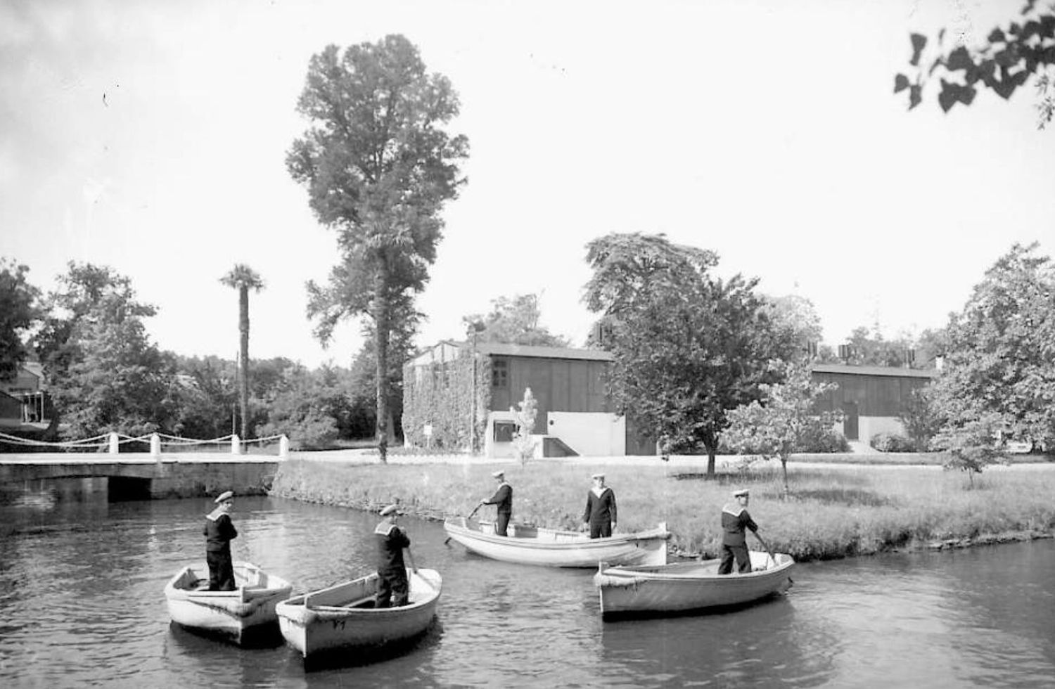 Black and white photo of people rowing boats on a river near a building surrounded by lush trees.