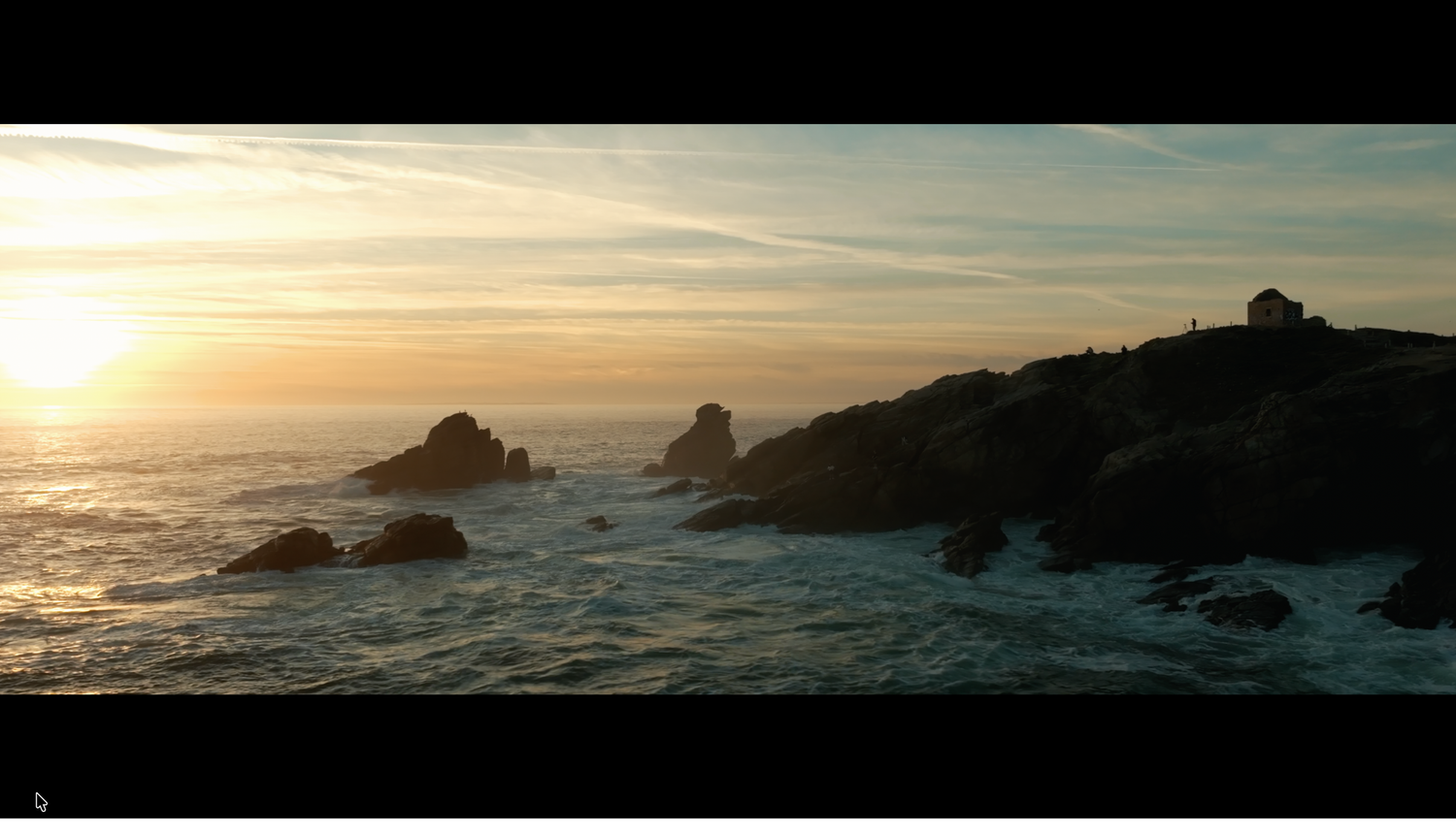 Dramatic sunset silhouetting rocky sea stacks and a small structure over misty ocean waters.