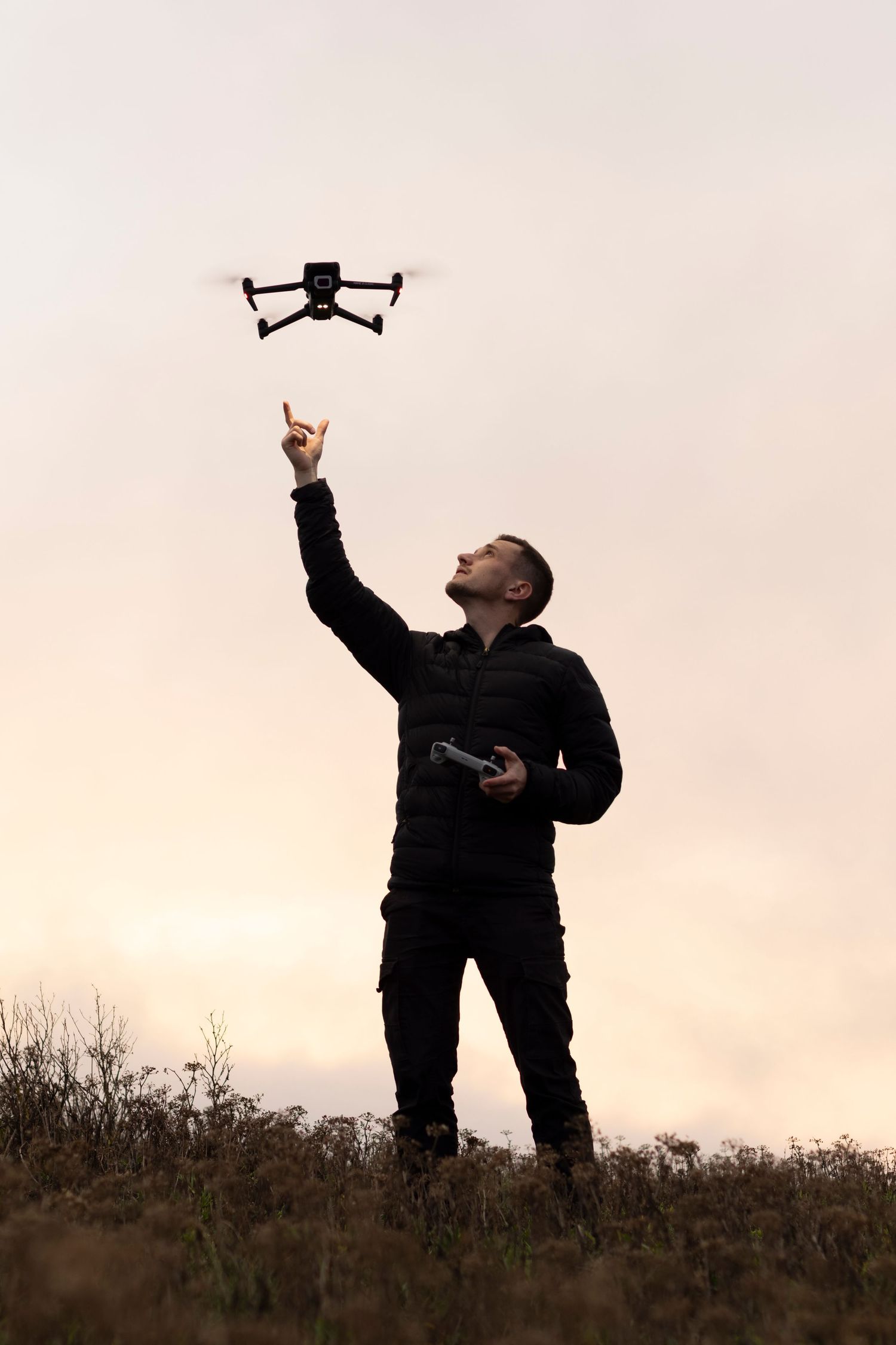 Man in black outfit pointing up at a small drone hovering above him in an open field at dusk.