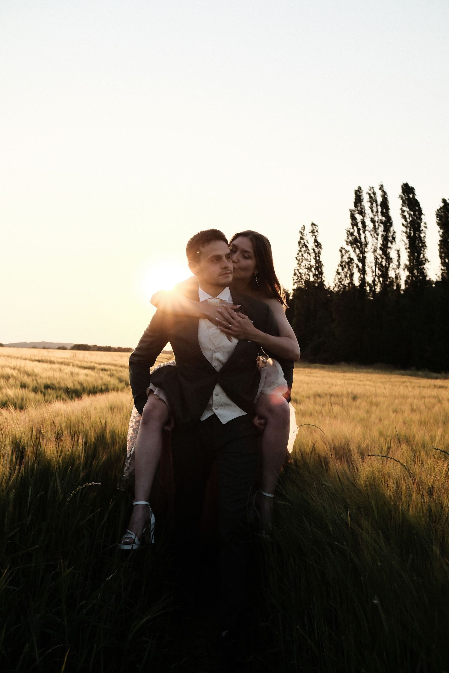 Groom carrying bride on his back through a sunlit wheat field at golden hour.