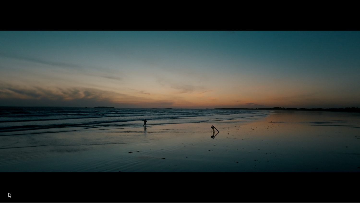 Two figures walking on a reflective beach at dusk with calm ocean waves in background.