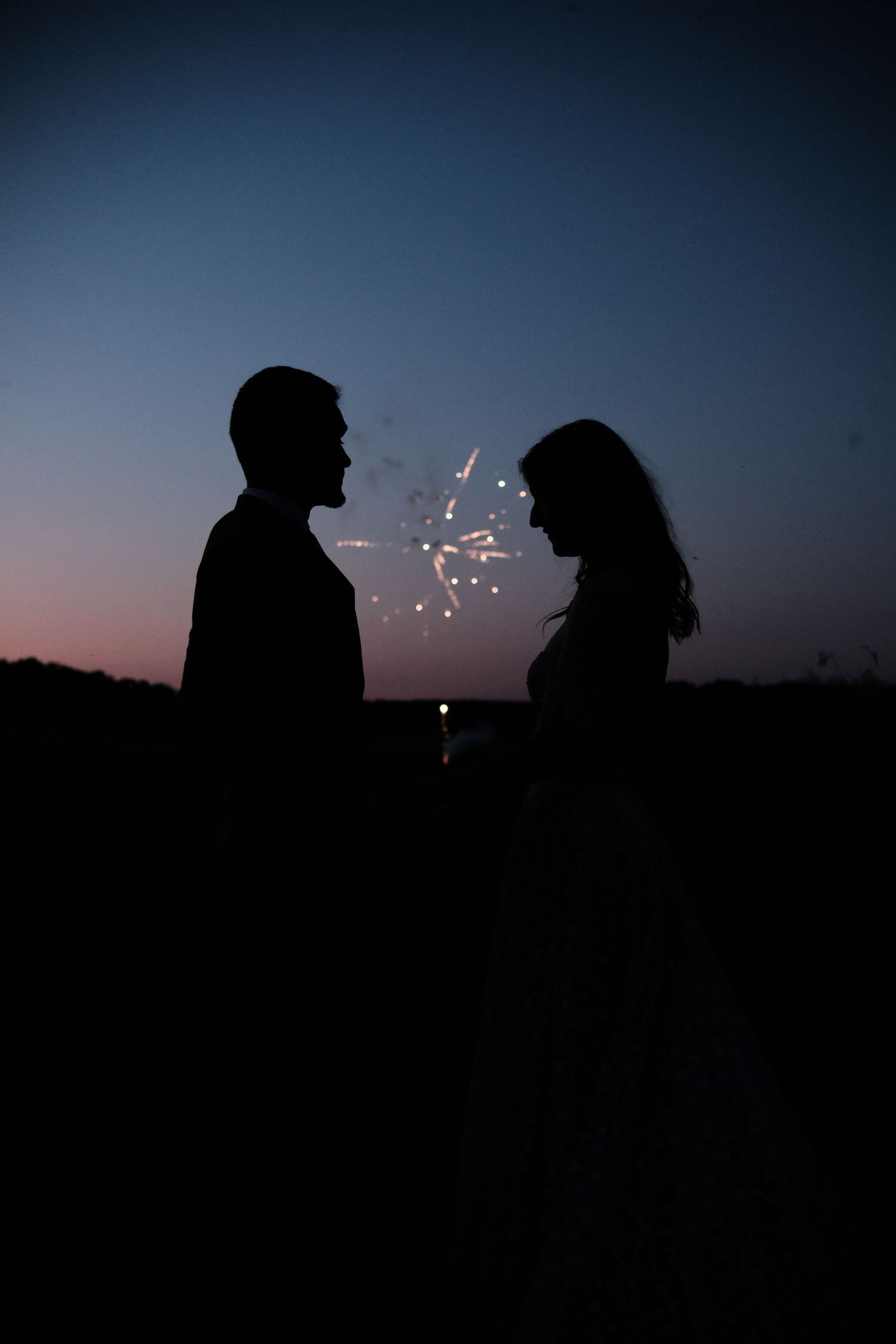 Silhouette of bride and groom facing each other with sparklers at twilight.