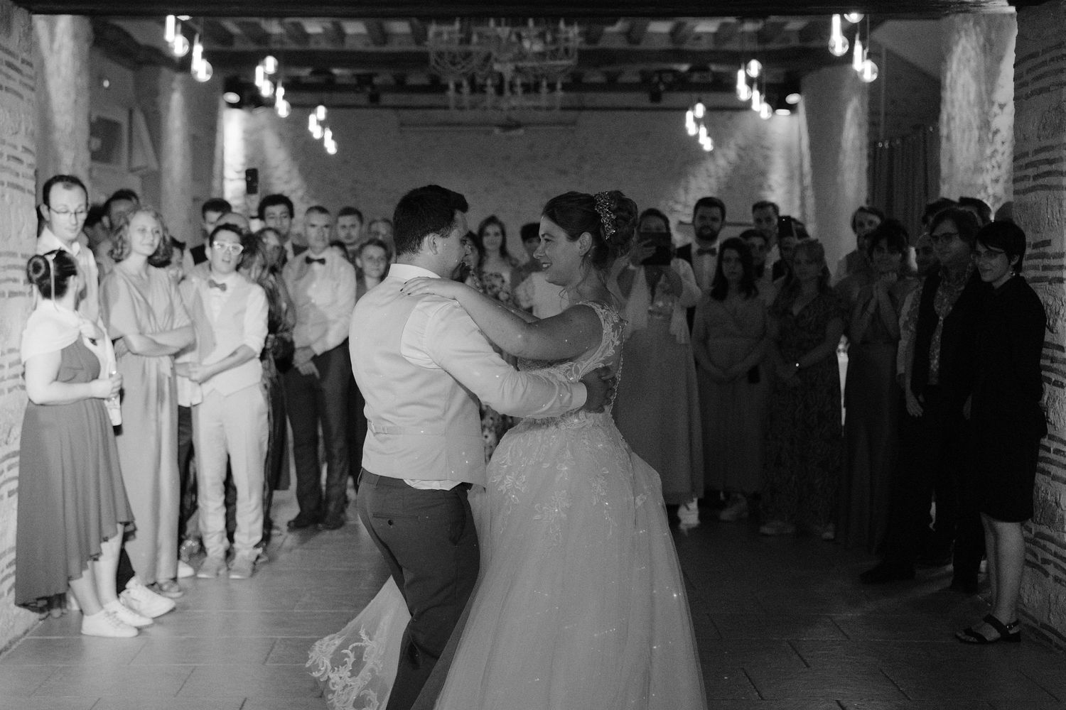 Black and white photo of bride and groom sharing first dance surrounded by wedding guests in rustic venue.