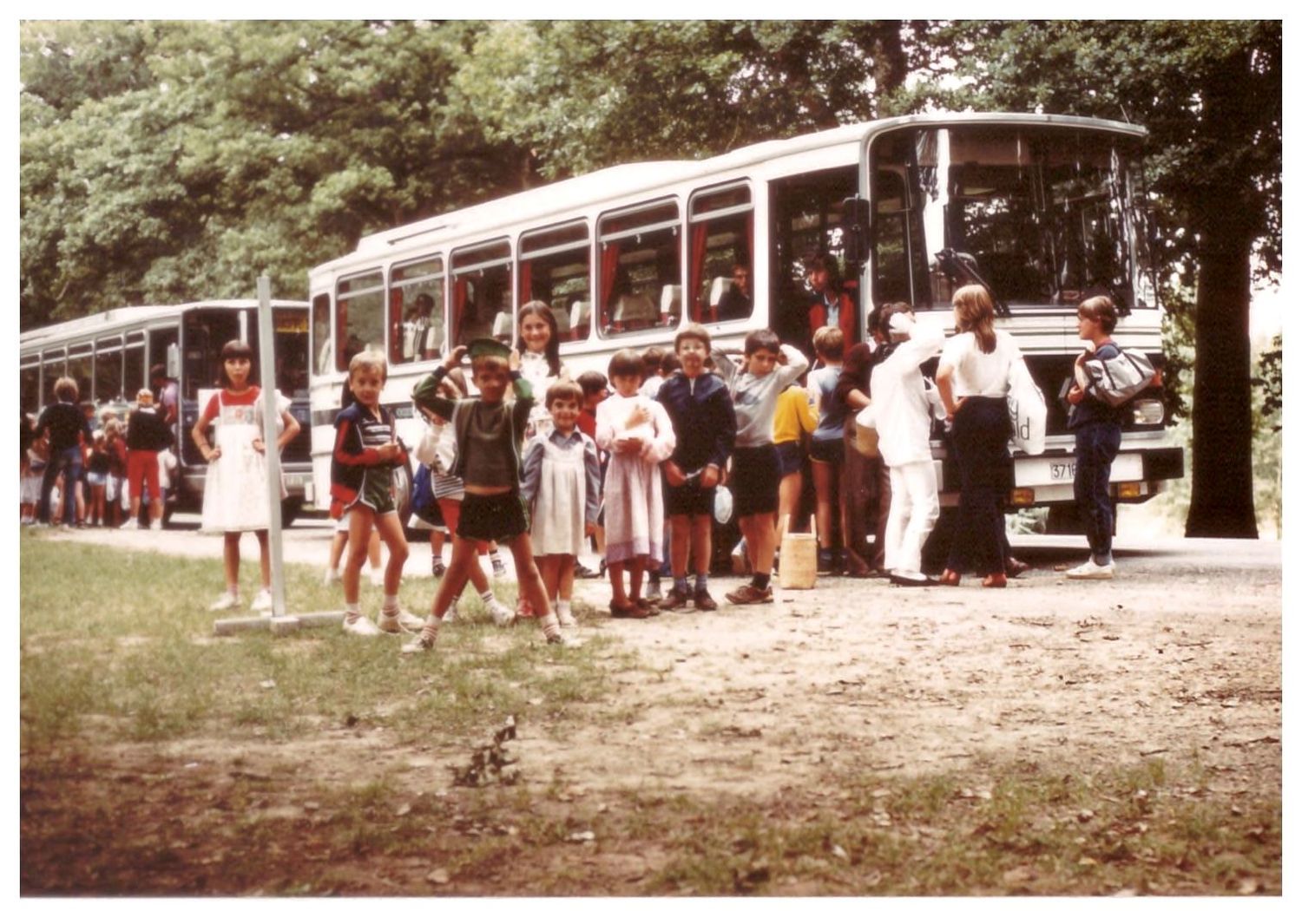 Children and adults gathered beside a large bus in a park setting in a color vintage photograph.