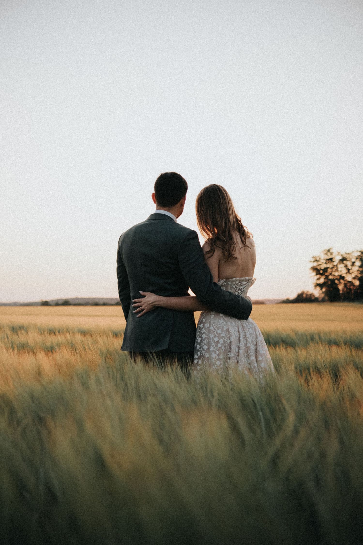 Wedding couple stands arm in arm in a lush green field at dusk.