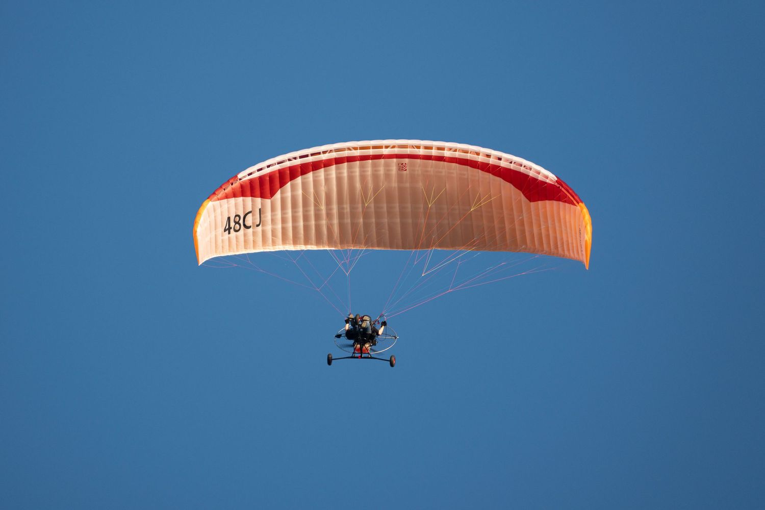 A motorized paraglider with an orange and red canopy soaring through a clear blue sky.