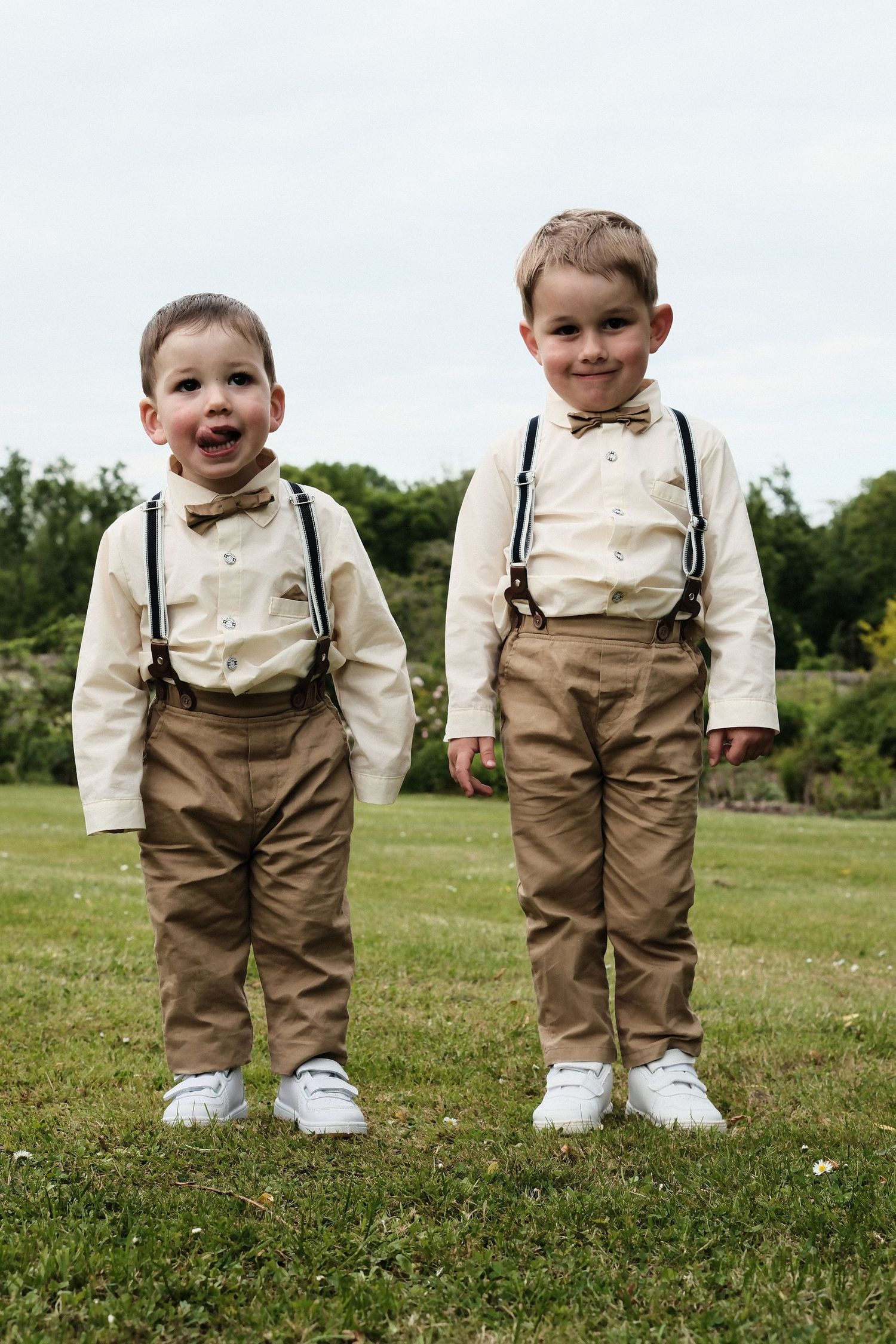 Two young boys dressed in matching beige suspender outfits with bow ties standing on a green lawn.
