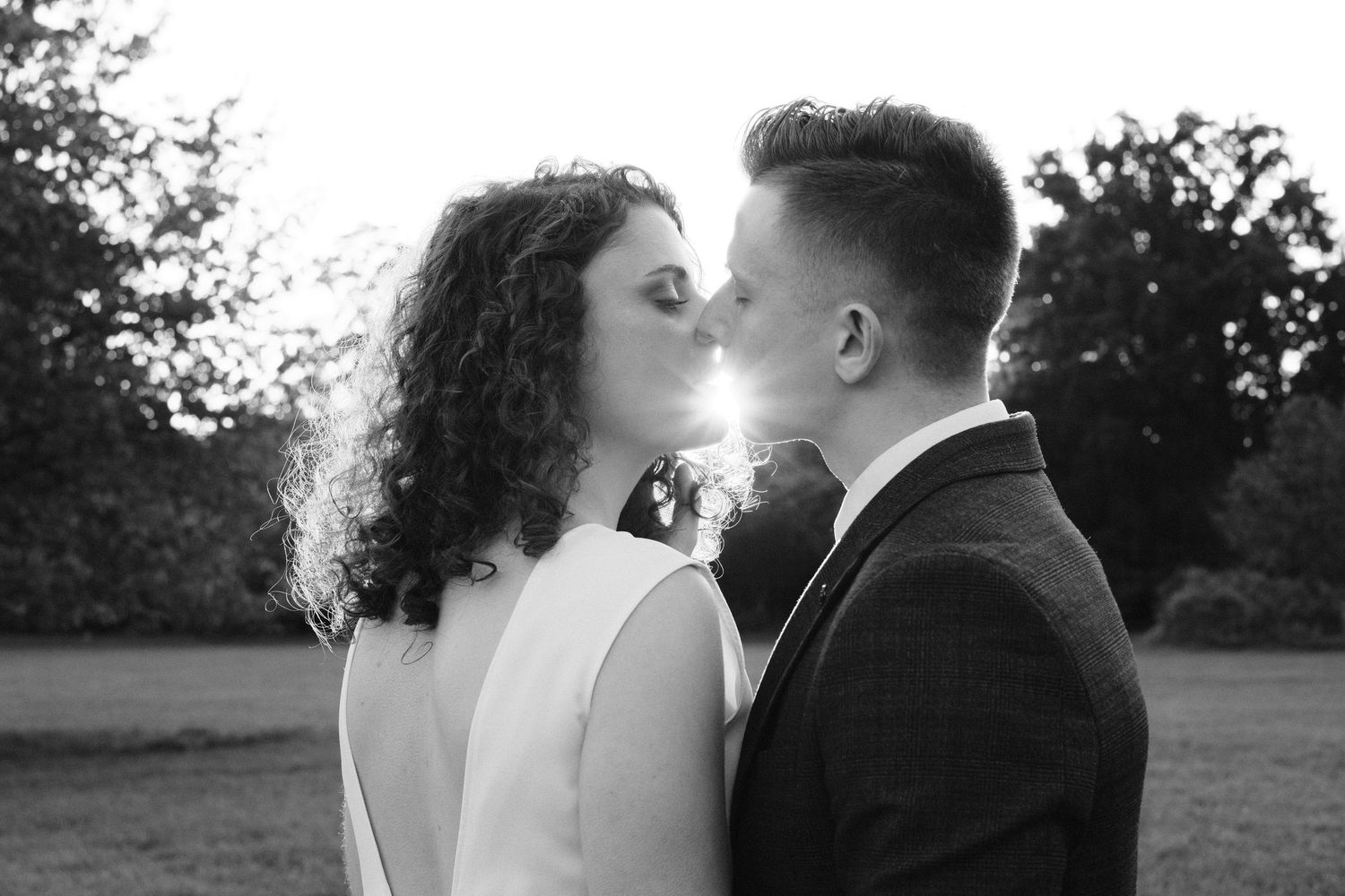 Black and white photo of a couple sharing a romantic kiss outdoors at sunset with trees in the background.