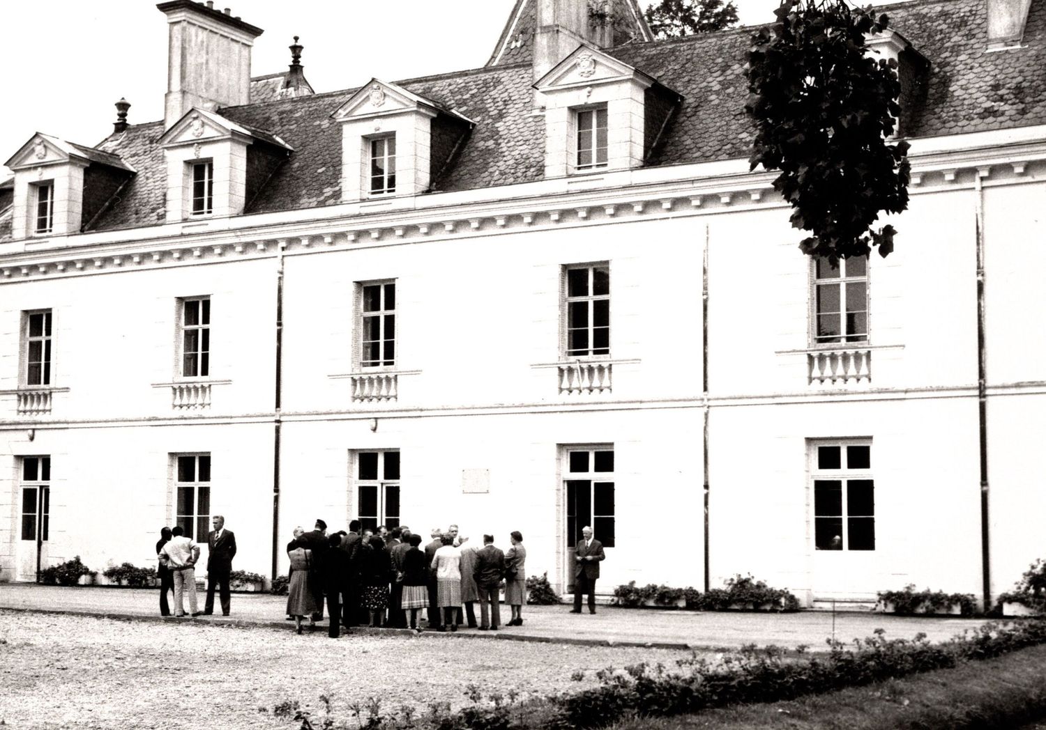 Black and white photo of a French chateau with mansard roof and group of people gathered outside.
