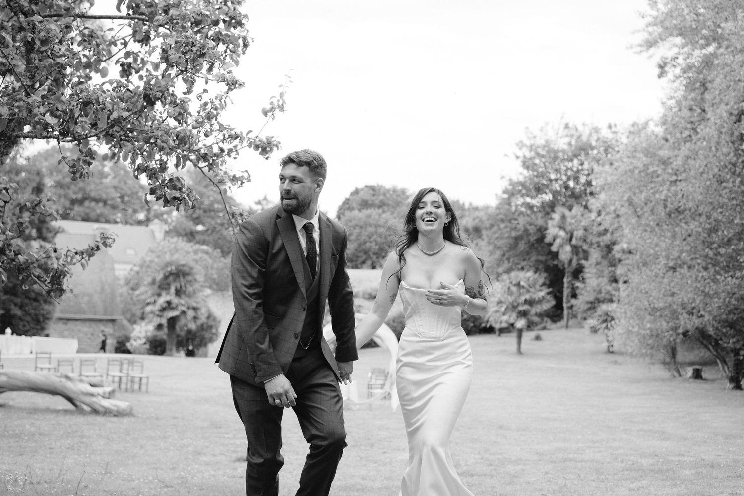 Black and white photo of a smiling bride in a satin gown and groom in a suit walking hand in hand outdoors.