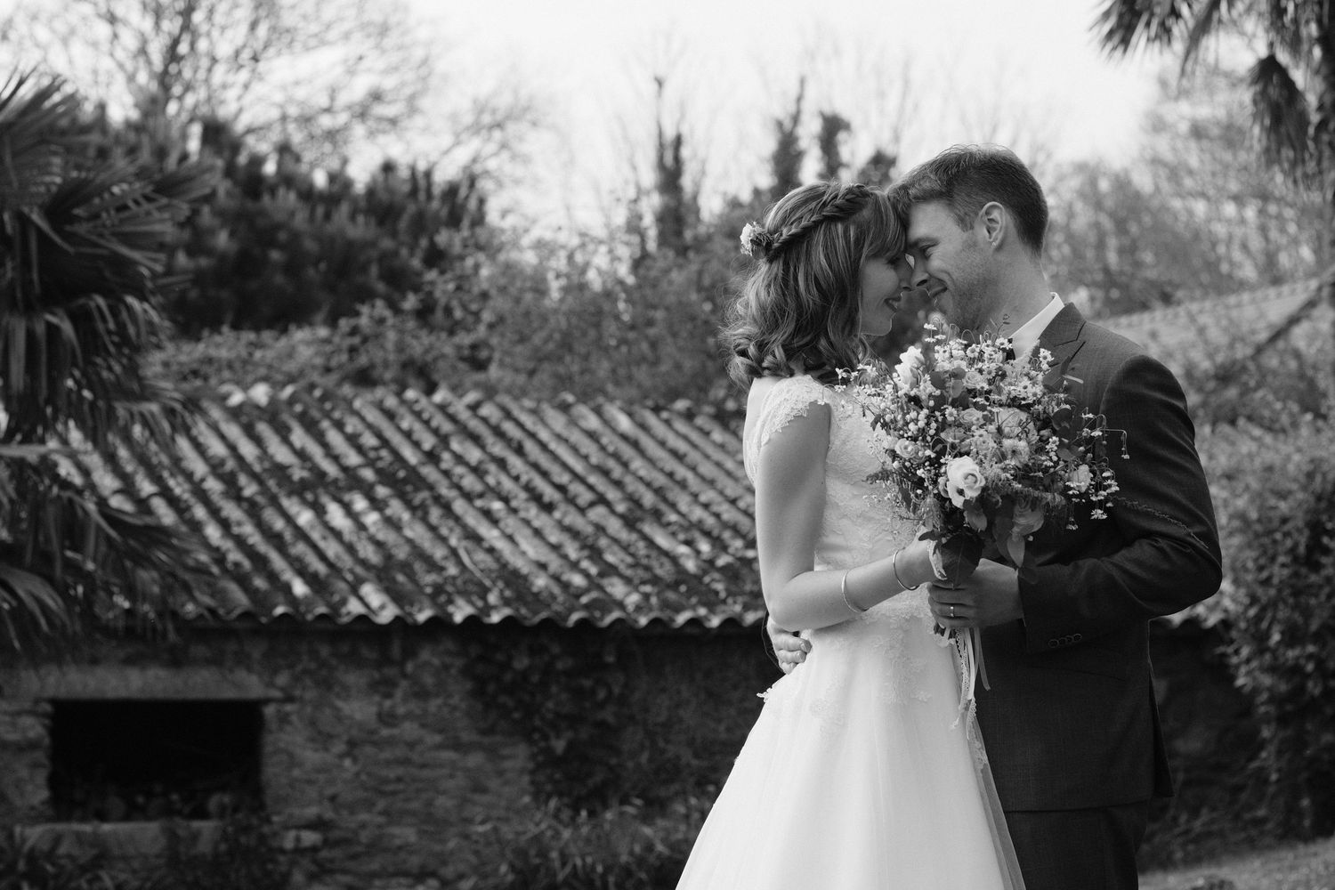 Black and white wedding photo of bride and groom touching foreheads, bride holding bouquet, rustic building background.