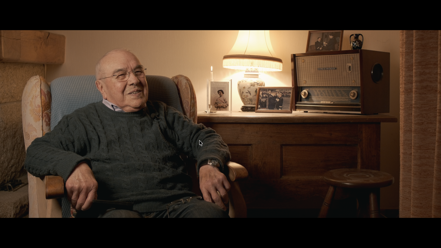 An elderly man smiles warmly while seated in a chair beside a vintage radio in a cozy room.