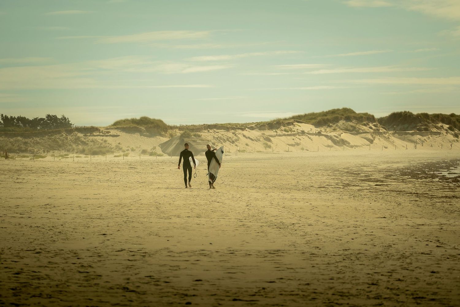 Two surfers carrying surfboards walk along a sandy beach toward coastal dunes under a hazy sky.