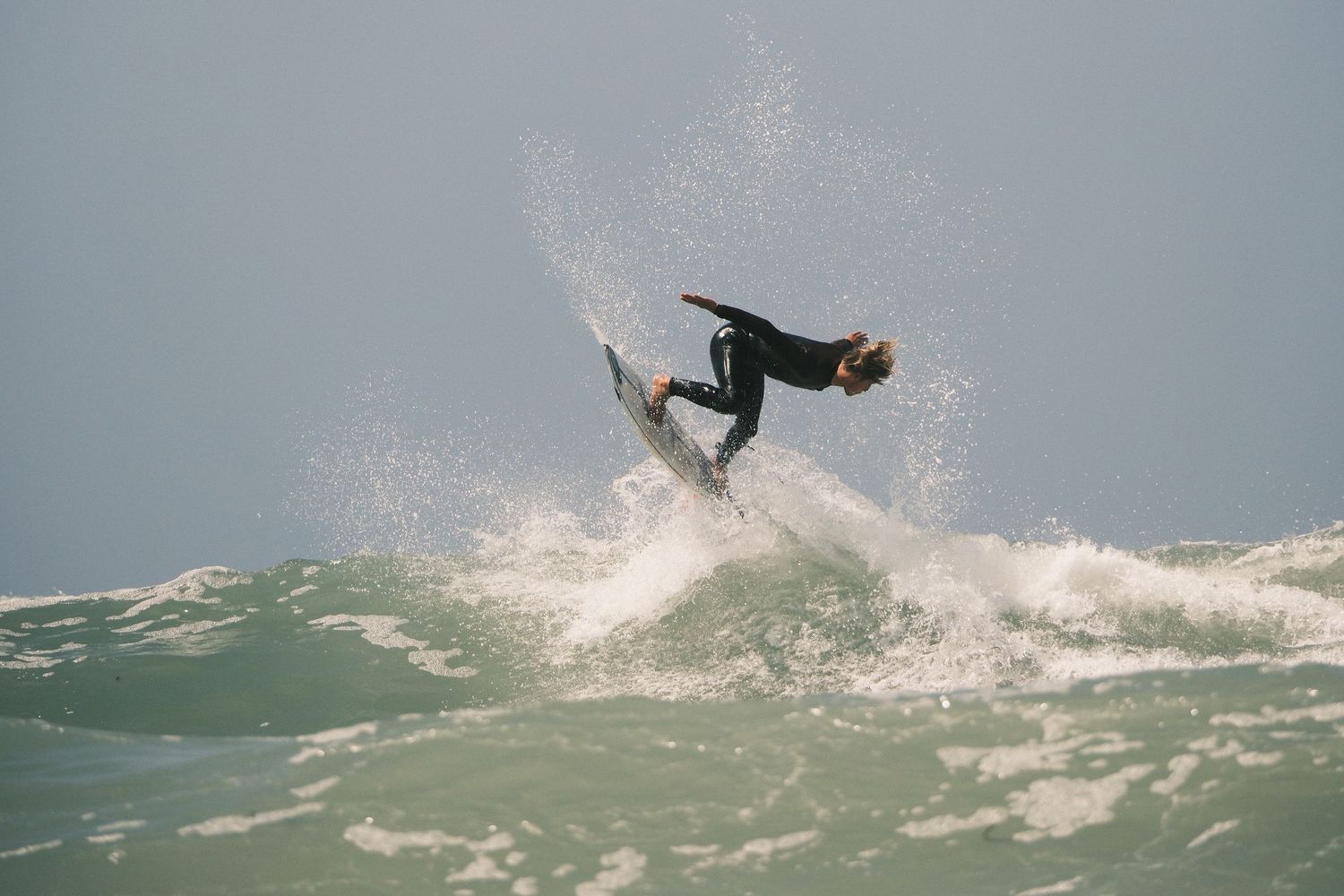 Surfer performing aerial trick on crashing ocean wave in misty sea conditions.