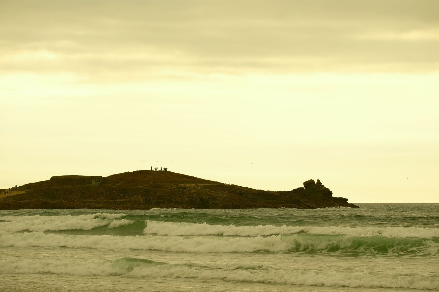Scenic ocean view at sunset with rocky island silhouette and rolling waves under golden cloudy sky.