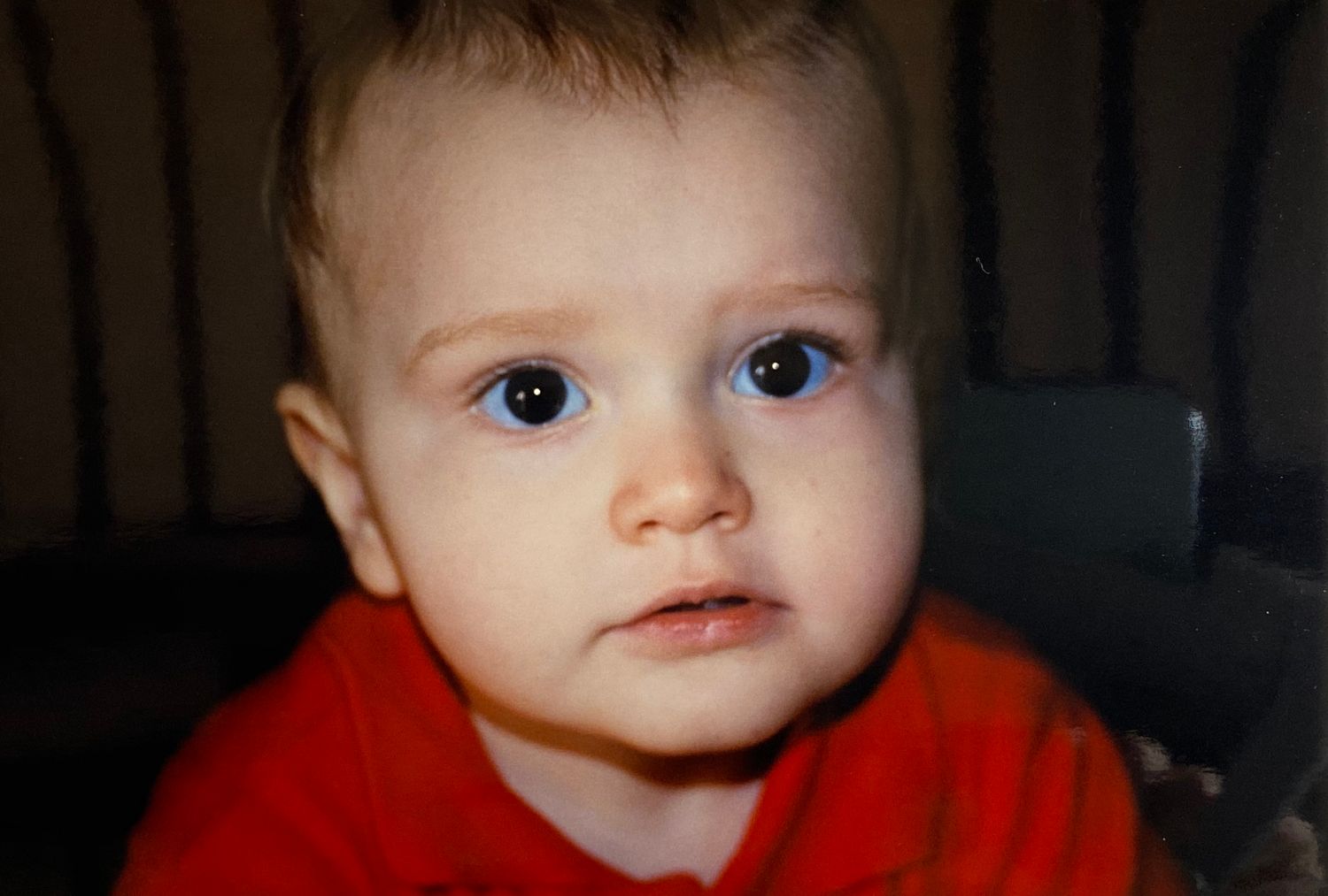 Close-up portrait of a blue-eyed baby in a red shirt with an innocent curious expression.
