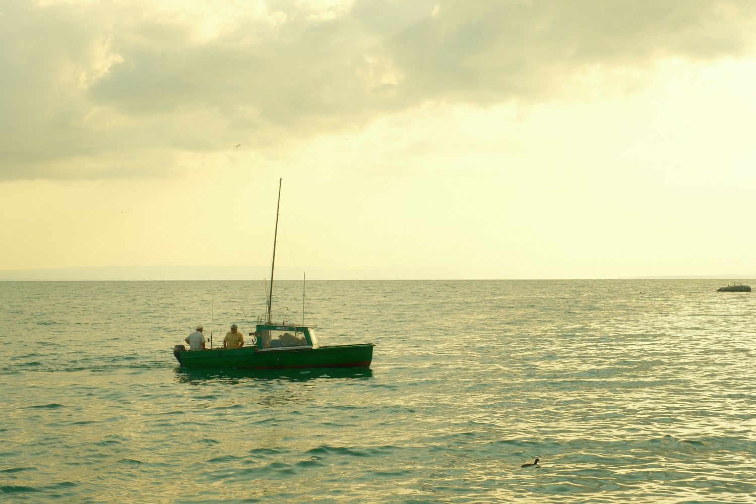 A small fishing boat with people on board sailing on calm ocean waters under a cloudy golden sunset sky.